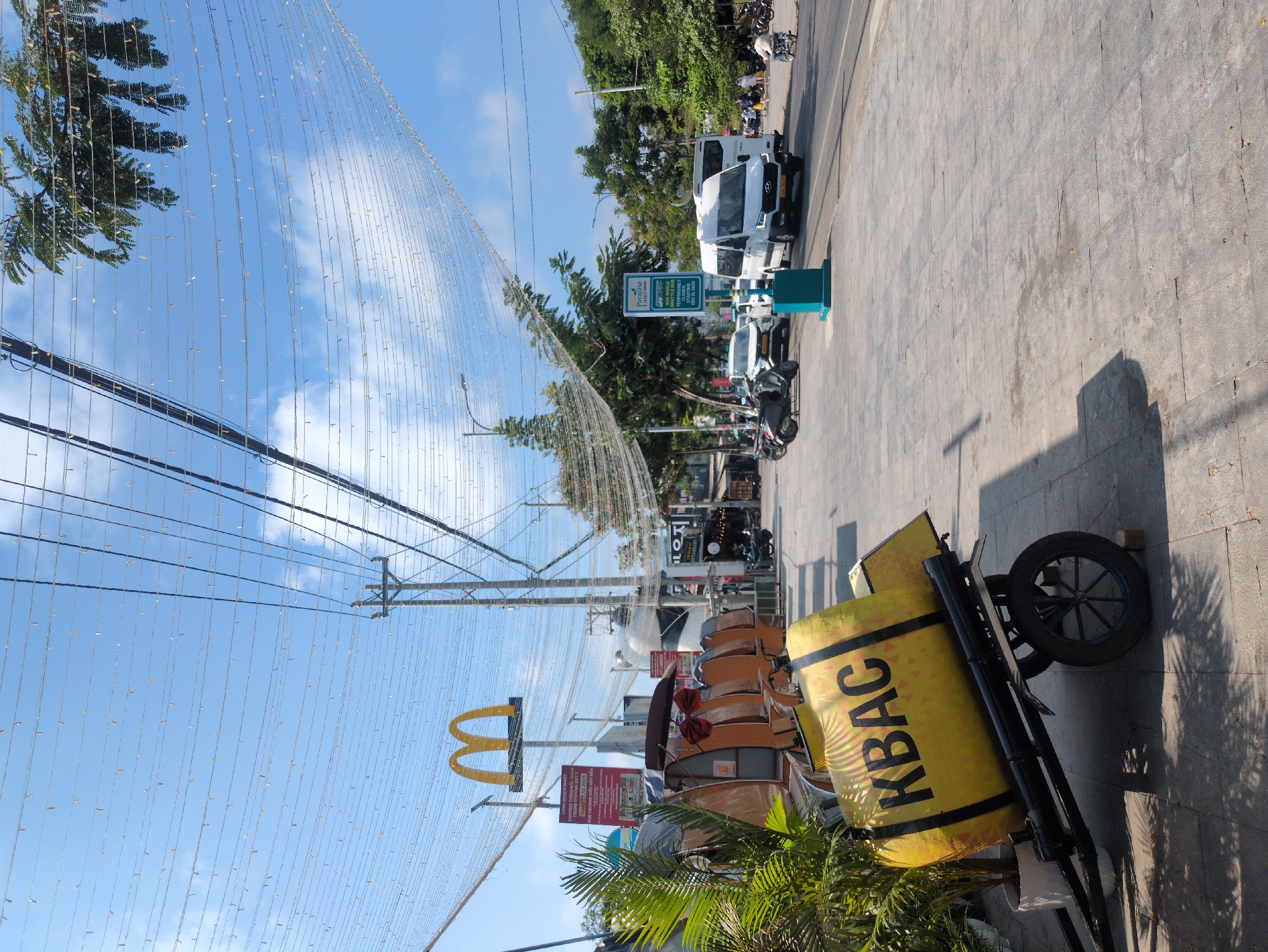 The image shows a street scene in Phú Quốc, Vietnam. The scene is on a sunny day. A yellow advertisement and a McDonald's sign are visible in the foreground. In the background, there is a street with parked vehicles and pedestrians. A sign indicates Paradise Island Shuttle Bus. Overhead, there are decorative strings of lights.