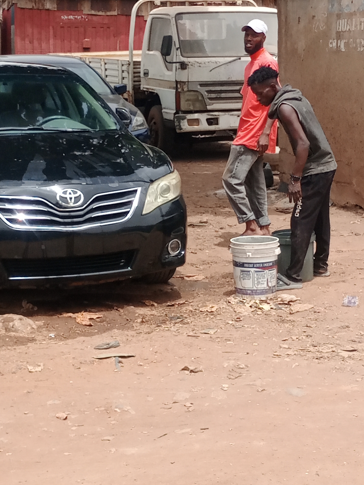 The image shows a street scene in Barden Yana, Nigeria. A black Toyota car is parked on a dirt road. Two men are standing nearby; one in a pink shirt and a white cap, and the other in a grey tank top and black pants. They appear to be washing the car. A white bucket and green container are on the ground near them. In the background, there is a white truck and a red building. The ground is a mix of dirt and debris.