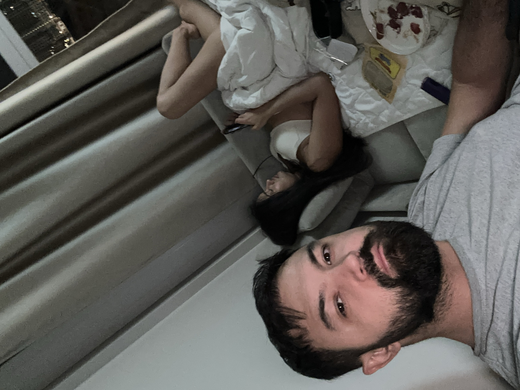 The image captures an intimate indoor scene featuring two individuals relaxing, likely during the evening in Khimki, Russia. The man, positioned prominently in the foreground, has dark hair and a beard, and appears to be taking a selfie. He is wearing a grey t-shirt and has a neutral to slightly content expression.
In the background, a woman is lying on a light-colored couch or bed, partially covered by a white duvet or blanket. She has dark hair, wears a white top, and is holding a smartphone, seemingly engaged with it. Next to her, on a flat surface, are a white plate with remnants of food, possibly berries and cream or yogurt, a small yellow and brown packet with some indistinct text, and a dark blue object that could be another smartphone or a remote control.
On the far left, pleated curtains in shades of grey suggest the presence of a window. The soft, indoor lighting implies that it is evening or nighttime. The overall atmosphere is casual and relaxed, depicting a moment of downtime.