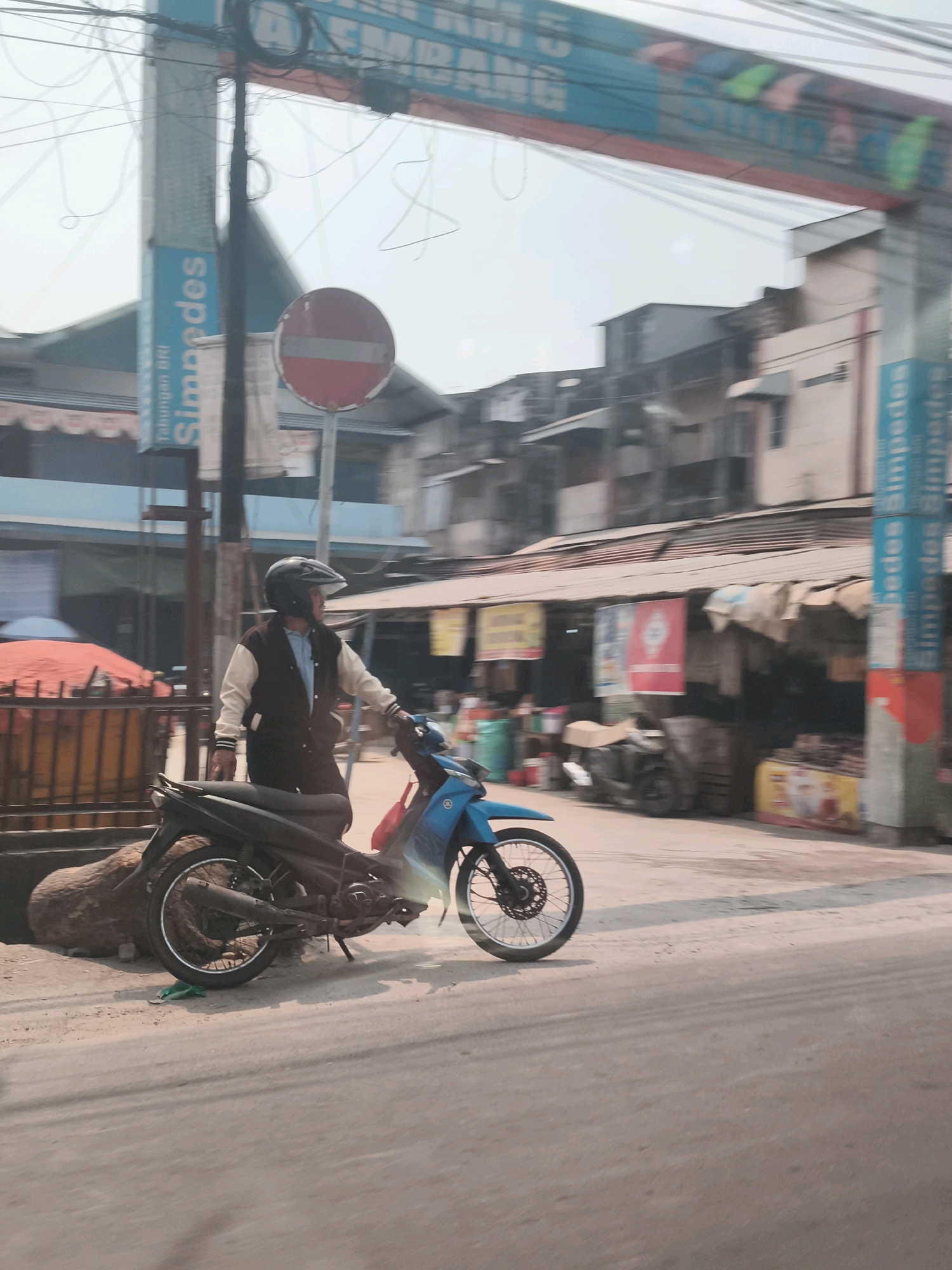 The image depicts a street scene in Palembang, Indonesia, specifically at the Simpang Tiga Empat Lawang intersection in Palembang. In the foreground, a man wearing a black helmet and a light blue shirt under a brown and white jacket is standing next to a blue motorcycle. The motorcycle is parked on the side of the road, with its front wheel facing left. Behind the motorcycle and the man, a street with buildings and shops lines the road. A red and white circular sign with a horizontal white bar is visible on a pole on the left side of the image. Vertical blue signs with white text "Simpedes" are also visible on poles. The sky is overcast.