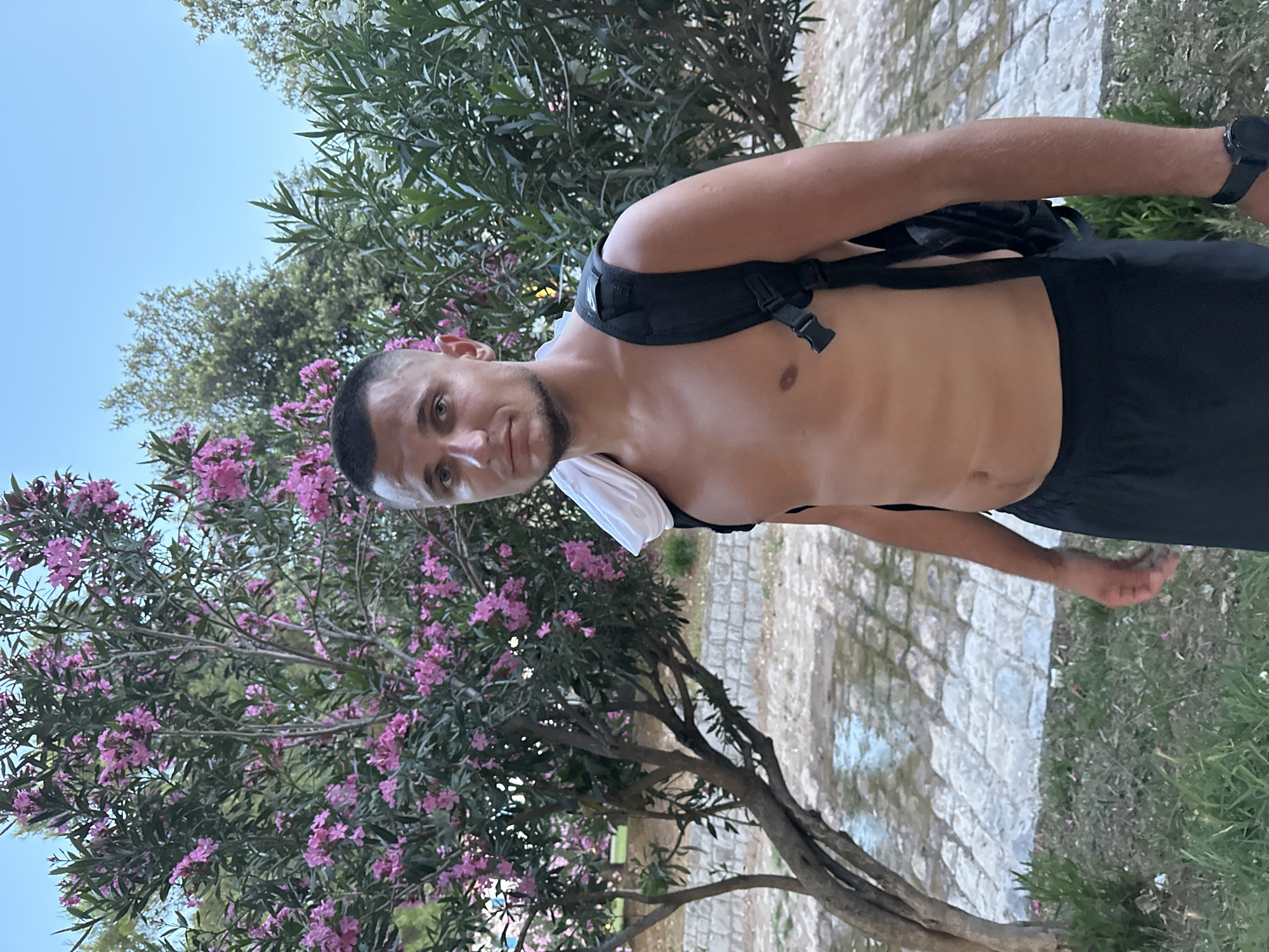 A shirtless young man with short brown hair stands outdoors, wearing a black backpack with a white towel draped over his shoulder. He is looking directly at the camera. He is wearing black shorts. In the background, there is a tree with pink flowers, lush green foliage, and a paved area with water on it. The scene appears to be in a park or garden area. The man is wearing a dark watch on his left wrist. The sky is clear and blue. The setting is likely Budva, Montenegro, given the presence of oleander flowers, which are common in the Mediterranean region, and the characteristic stone paving often found in coastal towns.