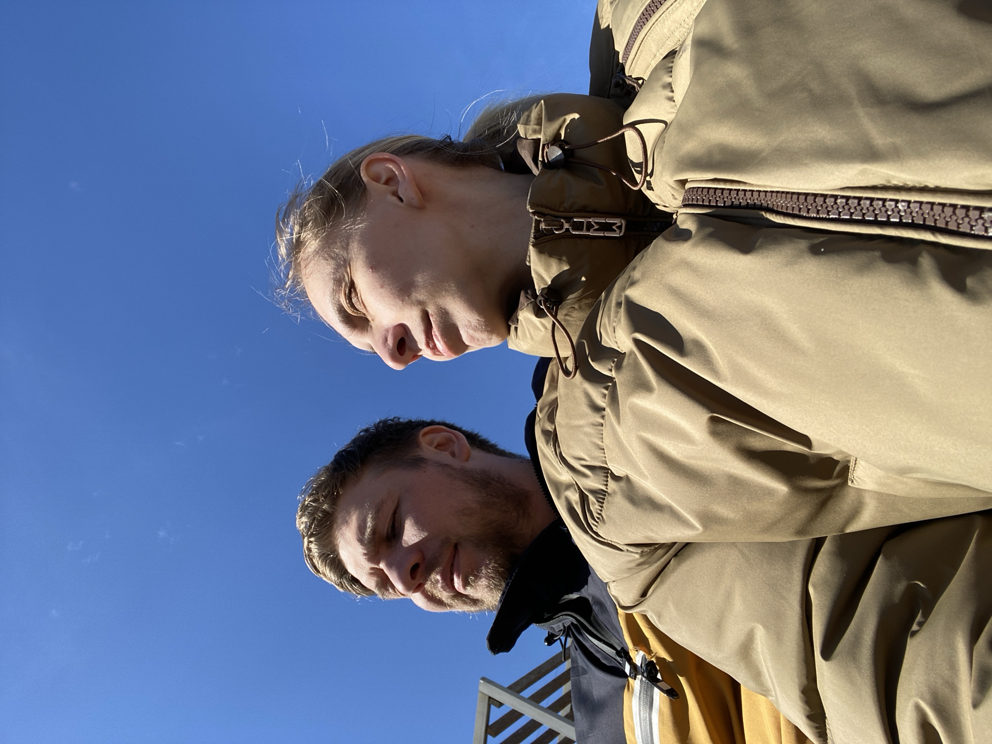 A low-angle, upward-facing shot captures two individuals against a bright blue sky. The person at the top of the frame is a fair-skinned woman with light blonde hair pulled back. She is wearing a khaki-colored puffy jacket. Below her is a man with fair skin, a beard, and short brown hair, also wearing a puffy jacket, parts of which are khaki, black, and yellow. A portion of what appears to be a wooden structure with parallel slats is visible at the bottom left. The location cannot be determined from the image.