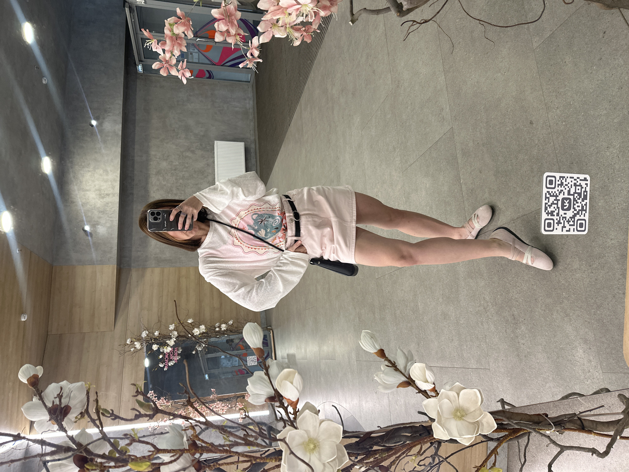 A young woman is taking a mirror selfie in a brightly lit, modern hallway. She is wearing a white long-sleeved shirt with a graphic design and a light pink mini-skirt with a black belt. She has a black crossbody bag over her shoulder and is wearing light pink flat shoes. The hallway has a concrete floor and walls, with a radiator visible on the wall. In the foreground, there are decorative branches with large white flowers. The mirror reflects the woman and the hallway, as well as a QR code sticker on the floor in front of her. The location is identified as the hallway of the "Olimpiysky" sports complex in Izhevsk, Russia.