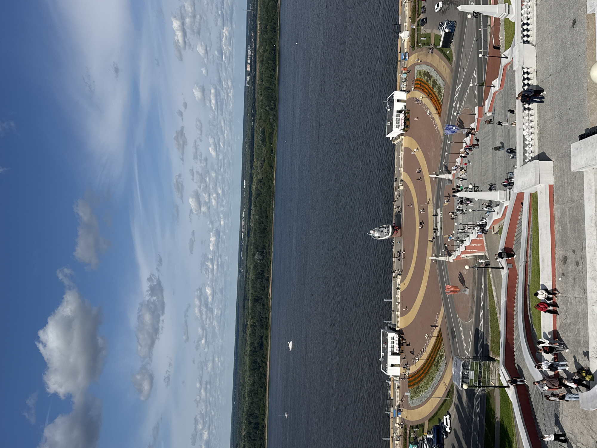 This image depicts a view of the Nizhny Novgorod Kremlin embankment and the Volga River. In the foreground, a wide set of stairs descends towards a circular plaza. People are scattered throughout the stairs and plaza, some sitting, others walking. The plaza itself is paved with a patterned surface and features flower beds, a white building with a red awning, and several lampposts.
Beyond the plaza, the dark blue waters of the Volga River stretch across the middle ground. Several boats are visible on the river, including a white motorboat and a larger white vessel docked on the embankment. On the far bank of the river, a dense green forest borders a cityscape with buildings. The sky above is a bright blue with scattered white clouds. The scene appears to be set on a bright, partly cloudy day.