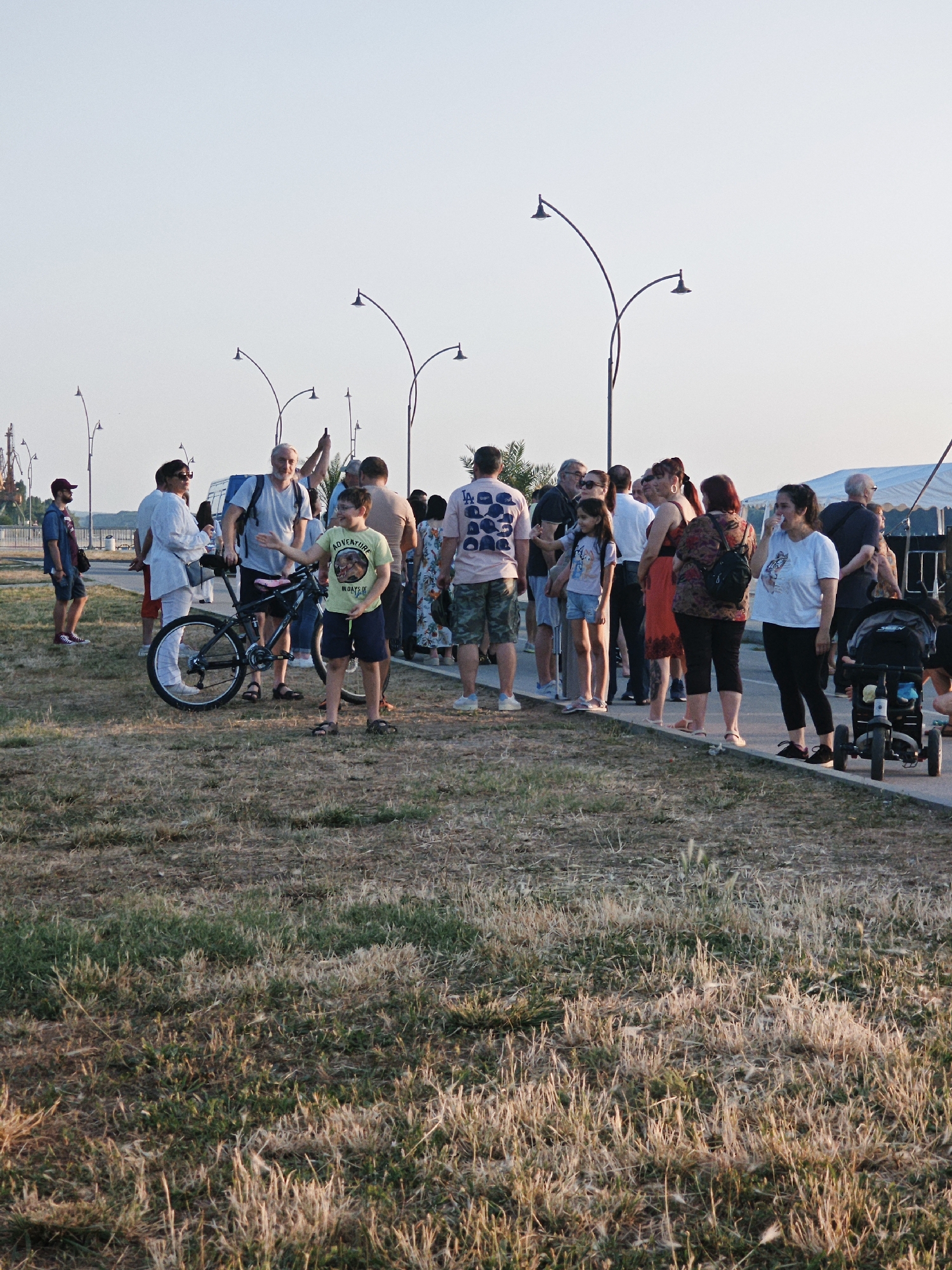 The image depicts a gathering of people on a paved walkway next to a grassy area. The scene appears to be outdoors, possibly near a waterfront or port, with industrial cranes visible in the background. The sky is clear and a pale blue. Several streetlights with curved poles line the walkway, and a tent structure is visible in the distance.
In the foreground, a young boy wearing a yellow t-shirt and dark shorts stands with a bicycle. He is gesturing with his right hand. Beside him, a man with a gray shirt and a backpack is looking at the boy. Several other people, of various ages and genders, are standing and conversing in groups along the walkway. Some are dressed casually, in t-shirts, shorts, and dresses.
A black baby stroller is parked on the right side of the walkway. The ground is covered with dry, yellowish grass and some patches of green vegetation. The overall impression is of a casual outdoor event or a leisurely stroll on a pleasant day.