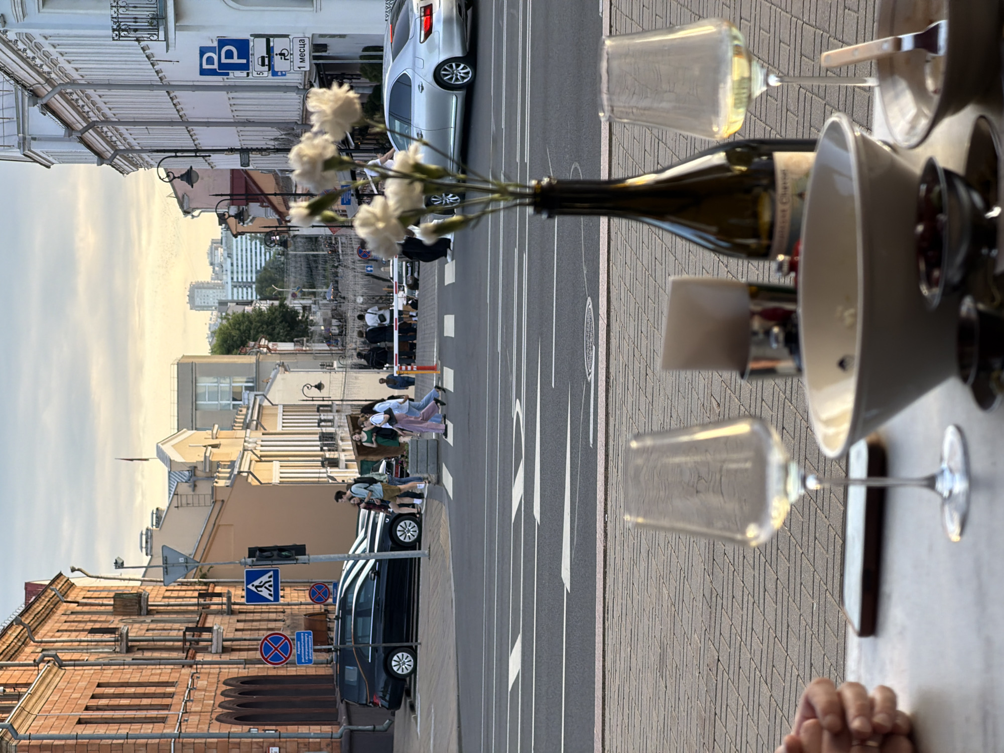 This is a street-level, medium-shot photograph taken at eye-level, likely from an outdoor seating area of a cafe or restaurant, looking towards a city street intersection. The setting is Minsk, Belarus, as indicated by the street signs.
In the foreground, on a table, are two empty wine glasses, a bottle of wine, a large white bowl, a small bowl with olives, and another partially visible bowl with a fork. The table surface is light gray, and the ground behind it is paved with tan and gray interlocking bricks.
The middle ground features a street intersection with pedestrian crosswalks. Several people are crossing the street: a group of three women and two men are walking across from left to right, and another person is walking across from right to left. A black hearse is parked on the left side of the street, and a silver sedan is driving away from the viewer on the right side of the street. Traffic lights and various street signs are visible, including a pedestrian crossing sign and parking signs.
The background shows a street lined with buildings. On the left, there is a historic brick building with arched windows. On the right, a white building with balconies and many windows lines the street. Further down the street, more buildings are visible, some with contemporary architecture. The sky is overcast with the warm glow of sunset or sunrise, and a few distant skyscrapers are visible on the horizon.
The overall impression is a candid, everyday scene in an urban environment, capturing a moment of people navigating a city street while others are dining.
