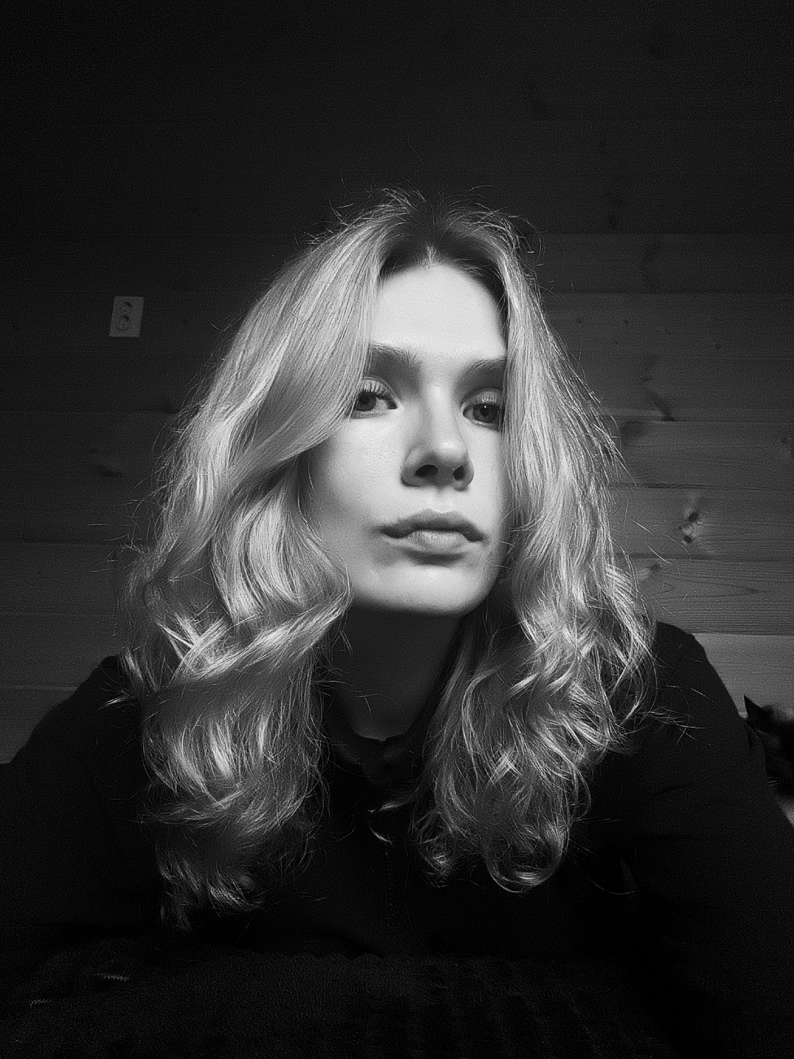 A black and white portrait captures a young person, likely female, from the chest up, looking directly at the viewer. She has long, wavy, light-colored hair that frames her face and extends over her shoulders, appearing thick and styled. Her facial features are distinct, with a neutral or slightly serious expression. She is wearing dark, possibly black, clothing that blends into the shadowed foreground, suggesting a top or hoodie.
The background is split between a dark, seemingly painted or heavily shadowed wall on the left, and a wooden plank wall on the right. On the dark wall, a white electrical outlet or light switch with two circular holes is visible near the upper left quadrant of the frame. The wooden wall consists of horizontal panels, adding a textural element to the scene.
The lighting is dramatic, characterized by strong contrasts, illuminating the subject's face and hair while casting much of the background and her clothing into deep shadow. This creates a moody and somewhat intimate atmosphere. The setting appears to be an indoor space, possibly a room in a home located in Русский Вожой, Russia. No specific activity beyond posing for the photograph is evident. There is no visible text in the image.