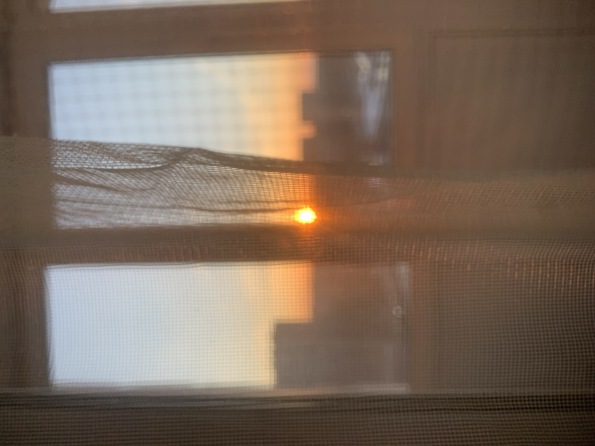 The image shows a view through a window covered by a sheer, patterned curtain. Outside the window, a blurred cityscape is visible, with the orange glow of the setting sun partially visible through the curtain. The architecture features indistinct building silhouettes. It is not possible to ascertain the specific location of Izhevsk, Russia, based on this image.