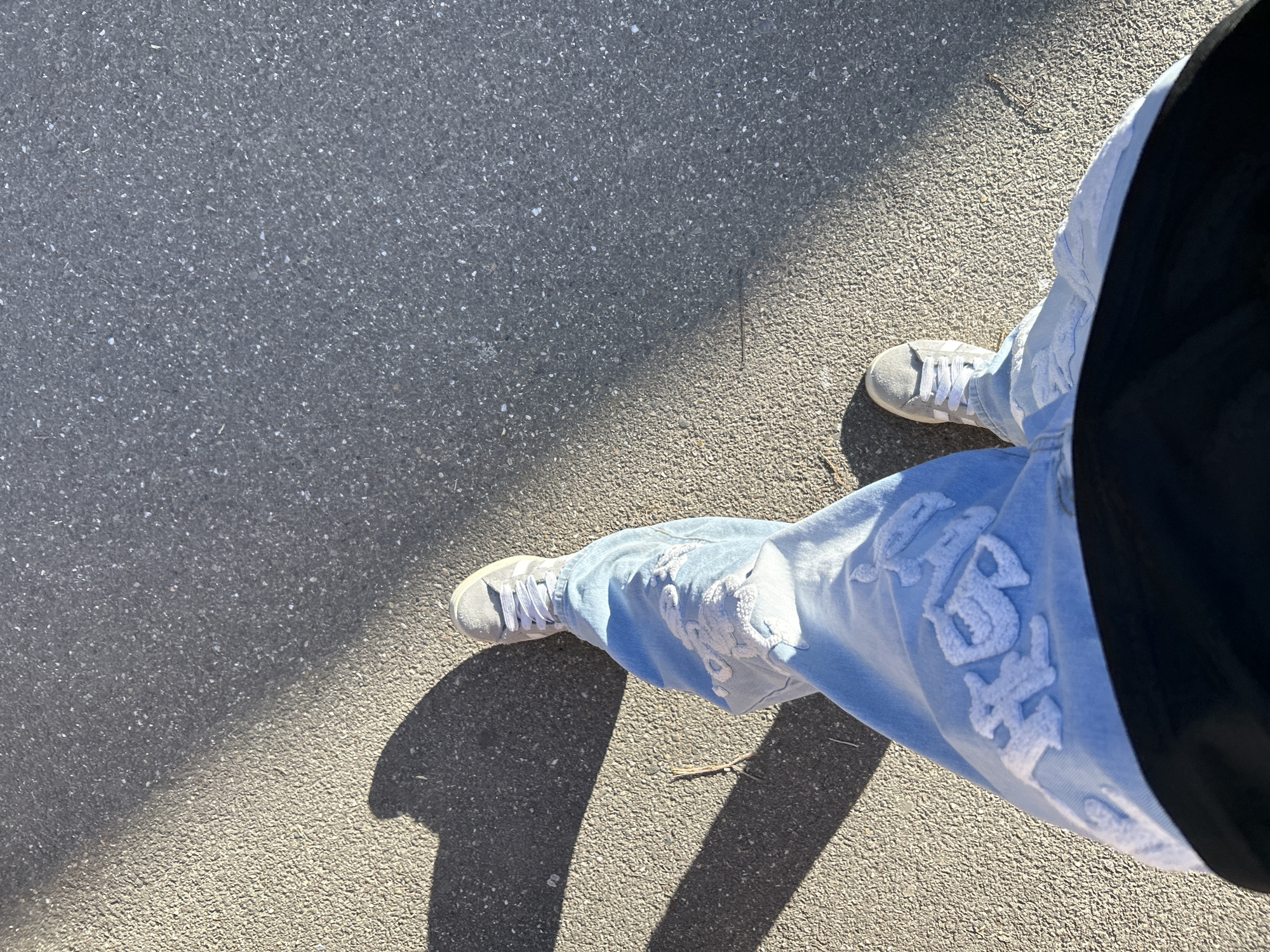 The image offers an overhead, downward-looking perspective of a person's lower body on an asphalt surface under bright sunlight in Strasbourg, France. The individual is wearing light blue, wide-leg jeans that feature prominent white, stylized text or embroidery, likely an appliqué design, on both legs. On the left leg, parts of three letters are visible; the middle letter is clearly a "G," and the last is an "H," while the first letter is partially obscured but appears to be an "S." This text has a raised, possibly puffy, and slightly distressed texture. The person's footwear consists of light grey sneakers with white laces and white stripes visible on the sides. A dark, likely black, garment is visible along the person's right side, suggesting a top or jacket.
The setting is an outdoor paved area, specifically dark grey asphalt, consistent with a street or sidewalk. Bright sunlight illuminates sections of the ground and the person's legs, creating distinct and sharp shadows across the scene. A large shadow occupies the upper-left quadrant of the frame, contrasting with the brightly lit areas. The clear delineation of light and shadow indicates a sunny daytime. The person appears to be standing or casually walking. No specific activity or emotion is discernible beyond this static pose.