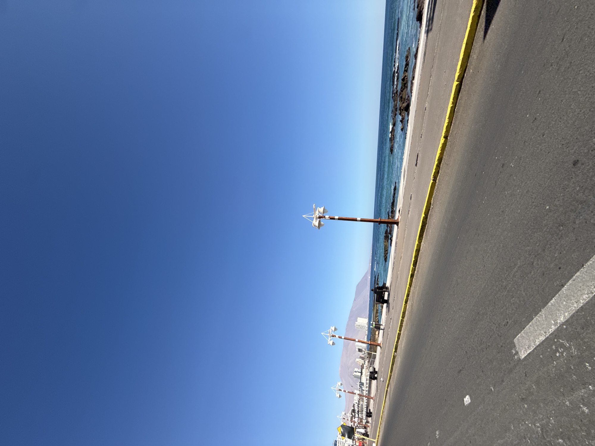 The image shows a coastal road in Iquique, Chile, likely along the Avenida Costanera. The scene is dominated by a clear, bright blue sky. In the foreground, a gray asphalt road with white lane markings is visible, with a yellow curb separating it from the sidewalk. Along the sidewalk, there are several lampposts with white light fixtures and brown and white striped poles. Two empty benches are placed along the sidewalk.
Further along the coast, there are buildings that appear to be part of the city, with some taller structures visible in the distance. The ocean is to the right of the road, with clear blue water and rocky outcrops. Waves are visible breaking against the rocks. In the background, a mountain range stretches across the horizon. The overall impression is of a sunny day in a coastal city.