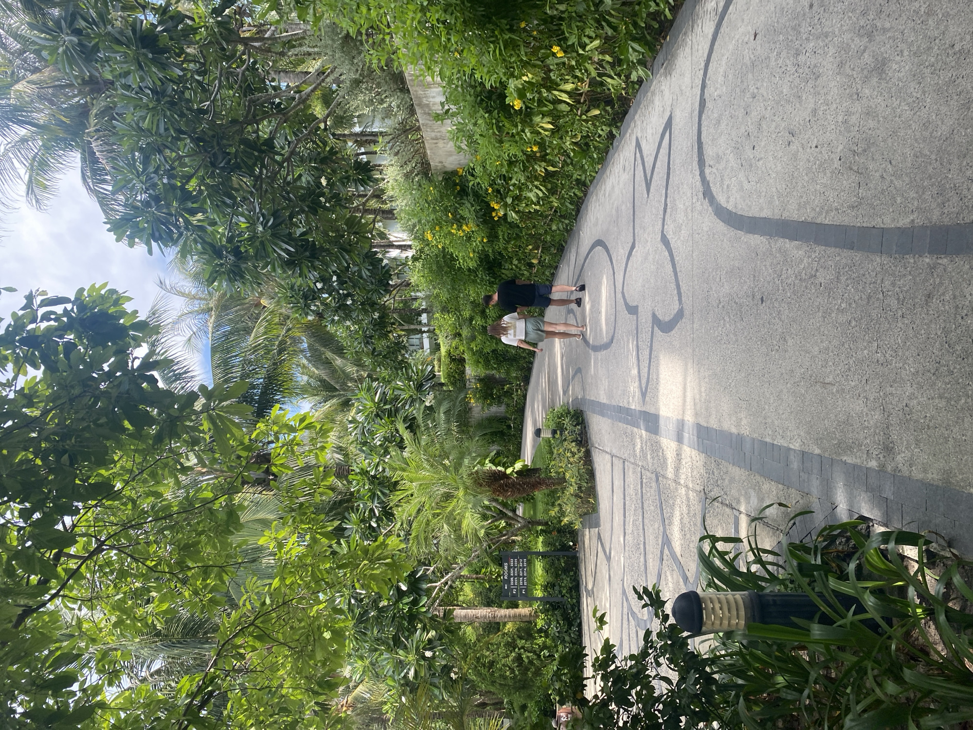 A photograph taken in Bac Cam Ranh Ward, Vietnam, depicts a paved walkway curving through lush vegetation. A man and a woman are walking away from the viewer on the path. The walkway is decorated with a repeating pattern of a stylized starfish-like design. Trees and tropical foliage line the path. In the middle ground, a sign with room numbers is partially visible amidst the greenery. A building is seen in the background. The image is taken at an oblique angle, possibly from a standing position, with a partial view of the sky through the foliage at the upper left.
