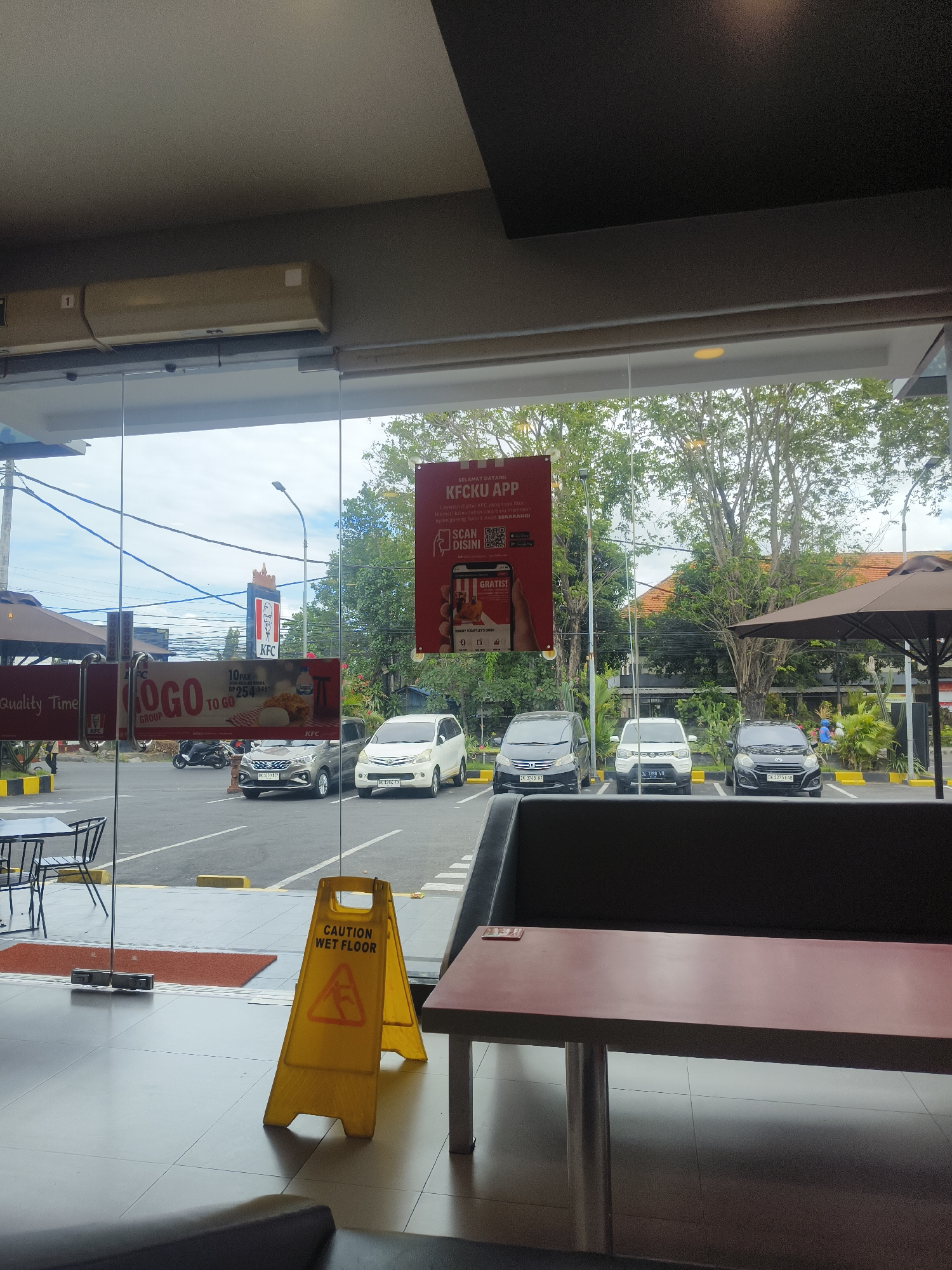 The image depicts the interior of a KFC restaurant in Denpasar, Indonesia, viewed through a glass partition. Through the glass, a parking lot and street scene are visible. The interior features tiled flooring, a dark-colored booth seat, and a reddish-brown table. A yellow "CAUTION WET FLOOR" sign with a wet floor symbol is prominently displayed on the tiled floor near the glass.
Outside, several cars are parked in designated spots. A white van is parked in the foreground, followed by two darker-colored cars. In the background, a KFC restaurant sign with the iconic Colonel Sanders logo is visible. Beyond the parking lot, trees and buildings form the urban landscape. The sky appears overcast.
A red banner promoting the "KFCKU APP" is hanging on the glass partition. Another banner is visible to the left, partially obscured, with text that appears to be "Quality Time" and "OGGO TO GO."
The lighting suggests it is daytime. No people are clearly visible in the interior. The overall scene is a typical view from inside a fast-food restaurant looking out onto its surroundings.