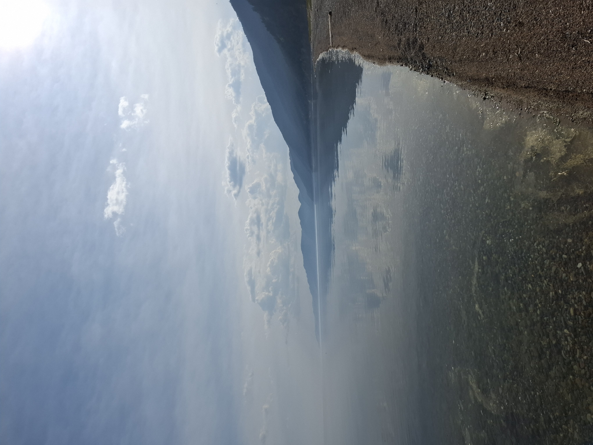 This media file captures a scenic view of a body of water, likely a lake, at the foot of a mountain range. The image is oriented vertically, with the sky and sun on the left, and the water and shore on the right. The sky is a bright blue with scattered white clouds, and the sun is partially visible, suggesting it is daytime, possibly late morning or early afternoon, with clear weather.
The mountain range forms a prominent silhouette against the sky, its dark slopes reflected in the calm water. The water's surface is smooth, creating a mirror-like reflection of the sky and mountains. Along the right edge of the image, a pebbly shore meets the water. The pebbles vary in size and color, and some are visible beneath the shallow water near the shore, indicating a clear water composition.
The overall mood is peaceful and serene, emphasizing the natural beauty of the location. There are no people or man-made objects prominently featured, focusing the attention on the landscape. The location is identified as Bugul'deika, Russia, which provides a geographical context for this tranquil natural setting.