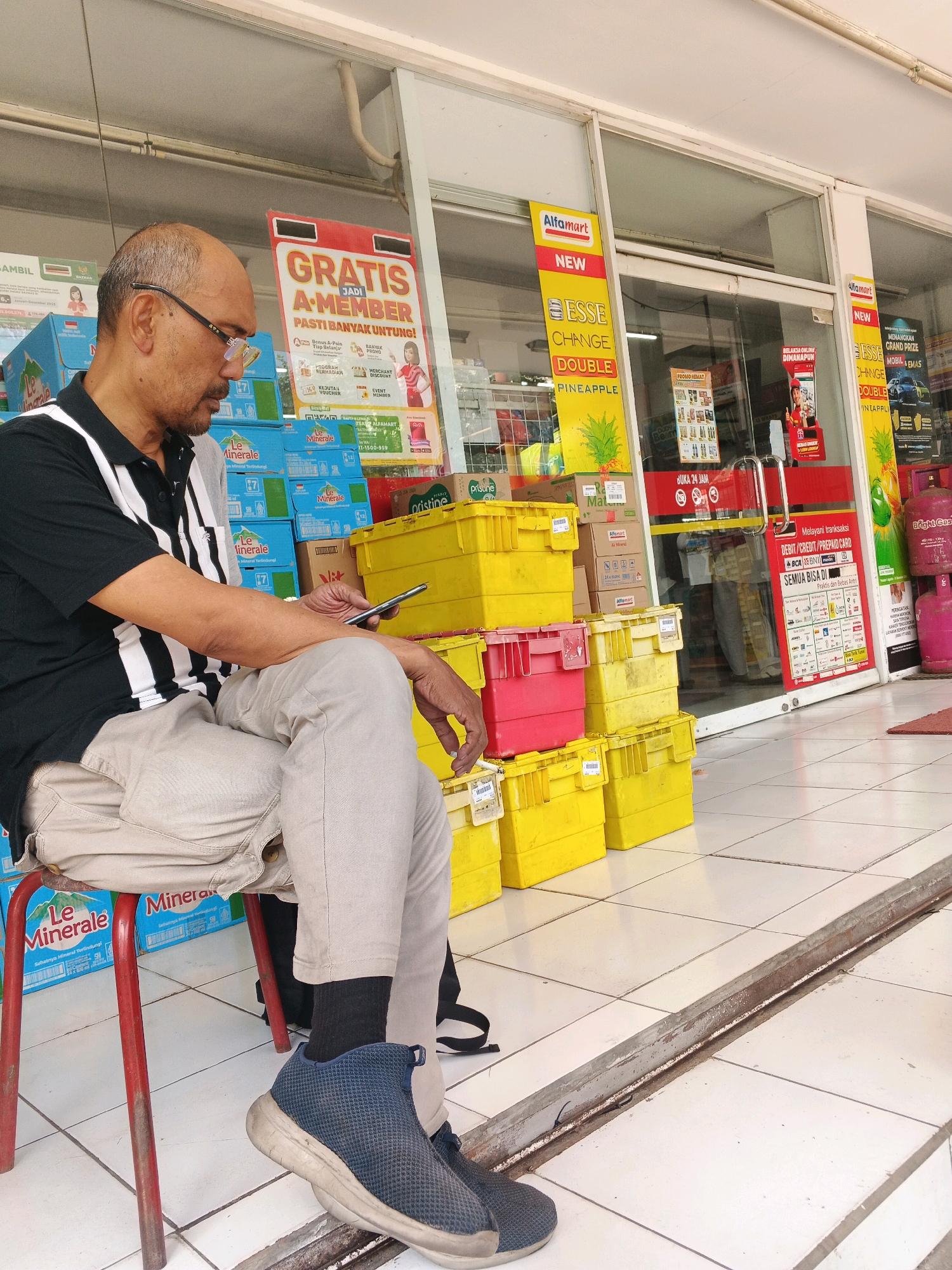 In the city of Kalirejo, Indonesia, an outdoor view of a convenience store reveals a bald man of middle age sitting on a red stool. He is wearing a black and white striped short-sleeved polo shirt, light-colored pants, dark socks, and blue athletic shoes. The man is looking down at his mobile phone, which he is holding in his right hand.
Behind the man, the store's exterior is visible, featuring large glass windows and doors. Stacks of bright yellow and red plastic storage bins are placed in front of the store, alongside stacks of bottled water. Promotional posters are visible on the store's windows, advertising memberships and product promotions. The ground is paved with white square tiles. The overall scene suggests a typical day at a local convenience store.