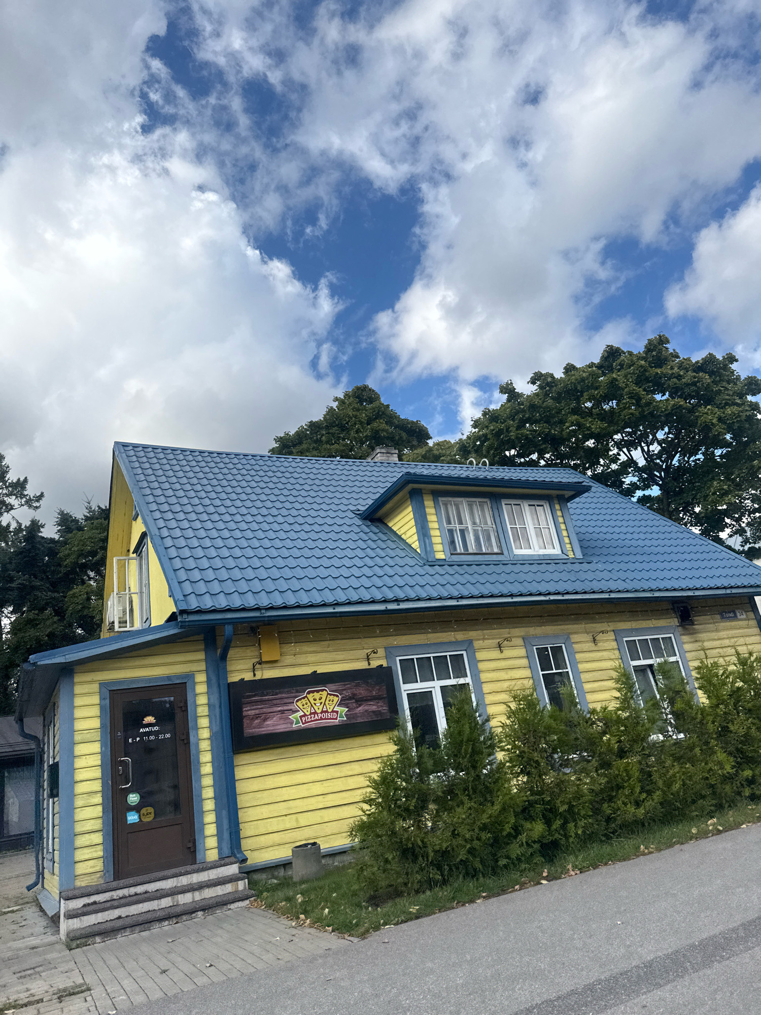 The image shows a bright yellow building with a blue metal roof, situated in Tallinn, Estonia. The building appears to be a commercial establishment, with a sign that reads "PIZZAPOISID" featuring cartoon pizza characters. A dark brown door with glass panels is visible, displaying opening hours from 11:00 to 22:00 on weekdays. There are several windows on the facade of the building, some with white frames and others with blue trim. A line of evergreen bushes runs along the front of the building, in front of some of the windows. The building is set against a backdrop of lush green trees and a partially cloudy blue sky. A paved walkway and a grey asphalt road are in the foreground. The overall scene suggests a casual dining or takeaway pizza place in an urban setting.