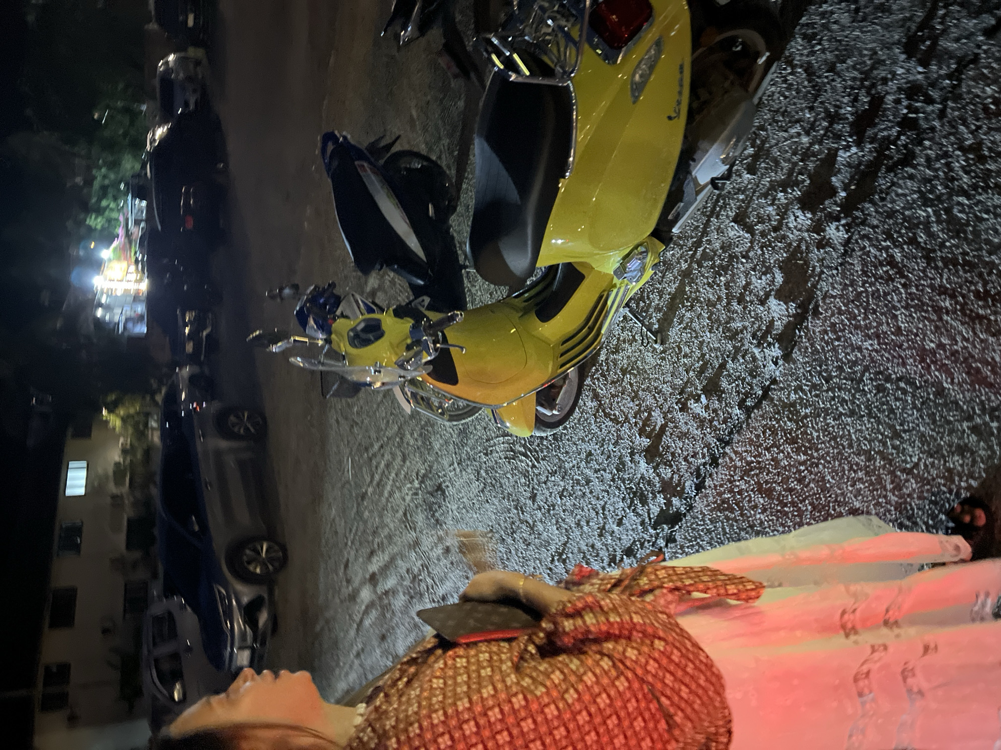 A person wearing a brown and red patterned garment is seen holding a black object near a bright yellow scooter, possibly a Vespa, parked on a textured, possibly wet, asphalt surface in what appears to be an urban setting. In the background, there are other parked vehicles and a building with lit windows. The scene is likely at night, given the lighting conditions, and the location is the city of Baan Bo Phut, Thailand.