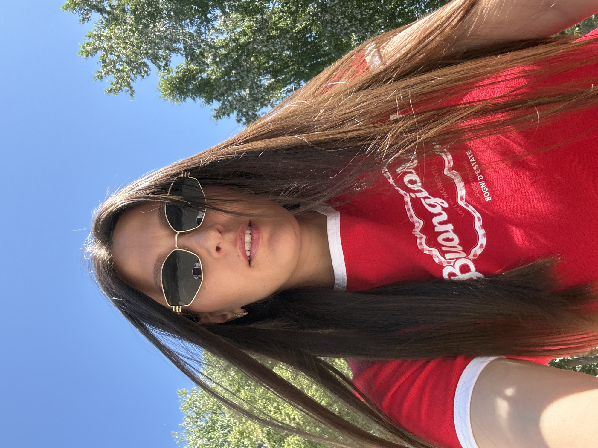 The image shows a close-up, low-angle selfie of a young woman with long, dark brown hair wearing sunglasses and a red t-shirt with white trim. The t-shirt has white text that appears to say "Buongiorno" and "Vivi il Momento, Gusto Italiano, Sogni d'Estate". She is outdoors, with a clear blue sky in the background and the tops of green trees visible on the right side. The lighting suggests it is daytime, possibly sunny. There is no indication of the city of Chaikovsky, Russia, or any other specific location.