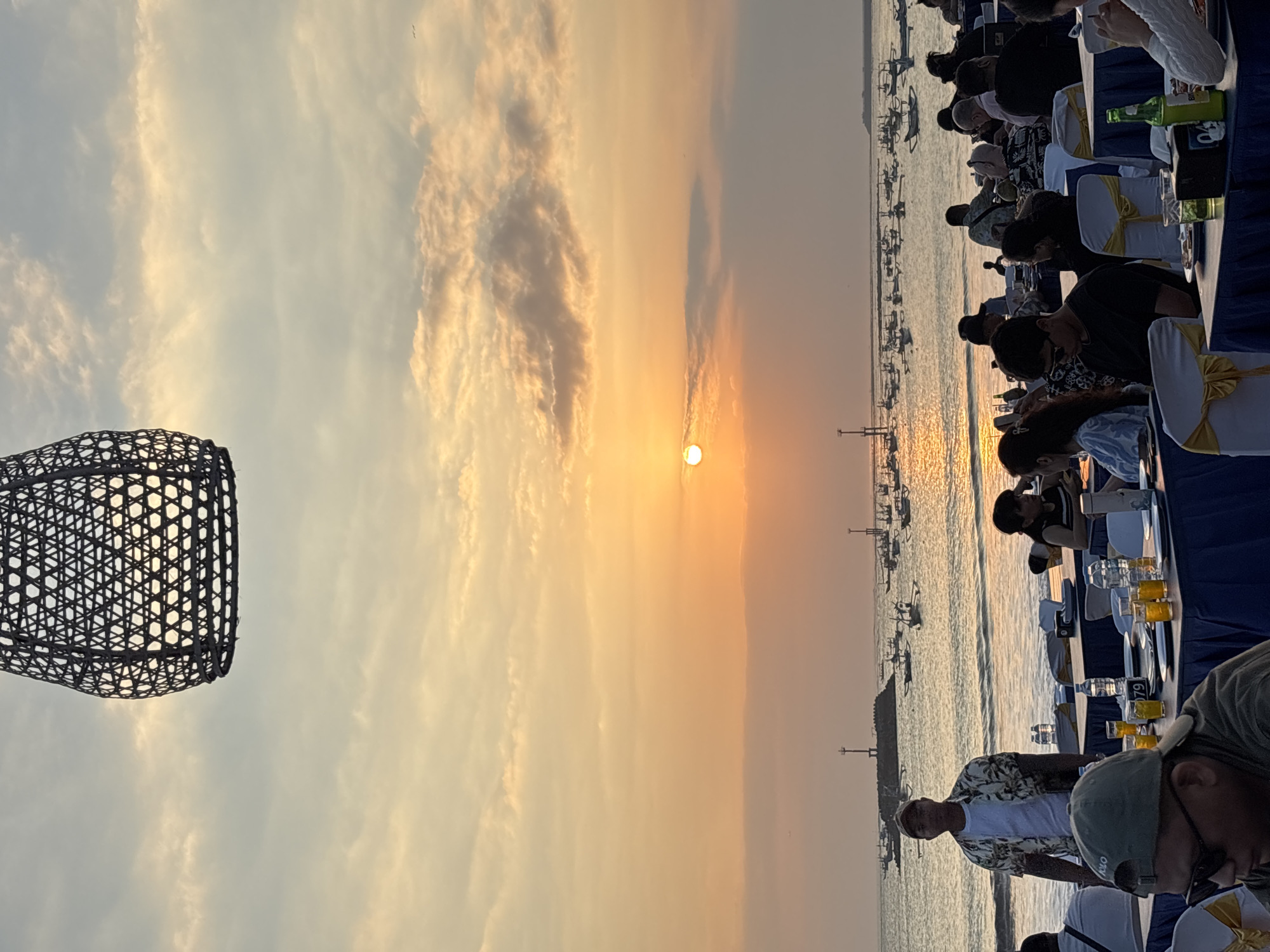 The image depicts a beachfront restaurant at sunset in Kuta, Indonesia. A large, woven hanging lamp is visible in the foreground, framing the sky. The sun is setting over the ocean, casting a warm, orange glow across the clouds and water. Numerous small fishing boats are scattered on the calm sea.
On the beach, tables are set up for dining, draped with blue tablecloths and adorned with yellow sashes. People are seated at the tables, some eating and conversing, while others are standing. A man in a floral shirt and a baseball cap stands near the tables, facing the camera. Another man in a baseball cap and sunglasses is seated at a table in the foreground. The scene suggests a relaxed evening gathering by the sea, with the natural beauty of the sunset as a backdrop.