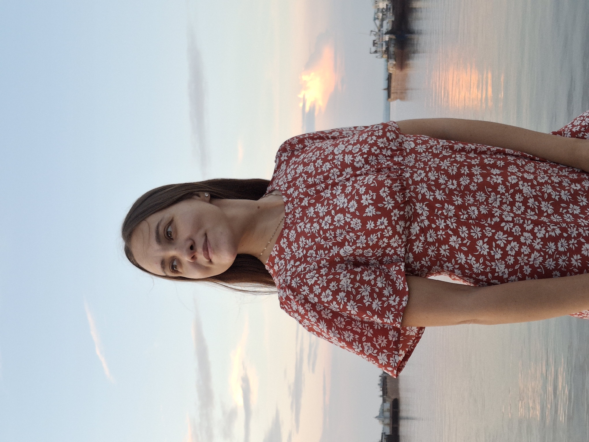 A young woman with long, brown hair stands facing left, looking up at the sky. She is wearing a red dress with a white floral pattern. The woman is positioned in the foreground, with a body of water behind her that reflects the sky. In the background, there are several boats and structures, suggesting a port or harbor area. The sky is a soft blue with hints of pink and orange from a sunset. The overall scene is serene and natural. The location is the embankment of the Kama River in Chaikovsky, Russia.