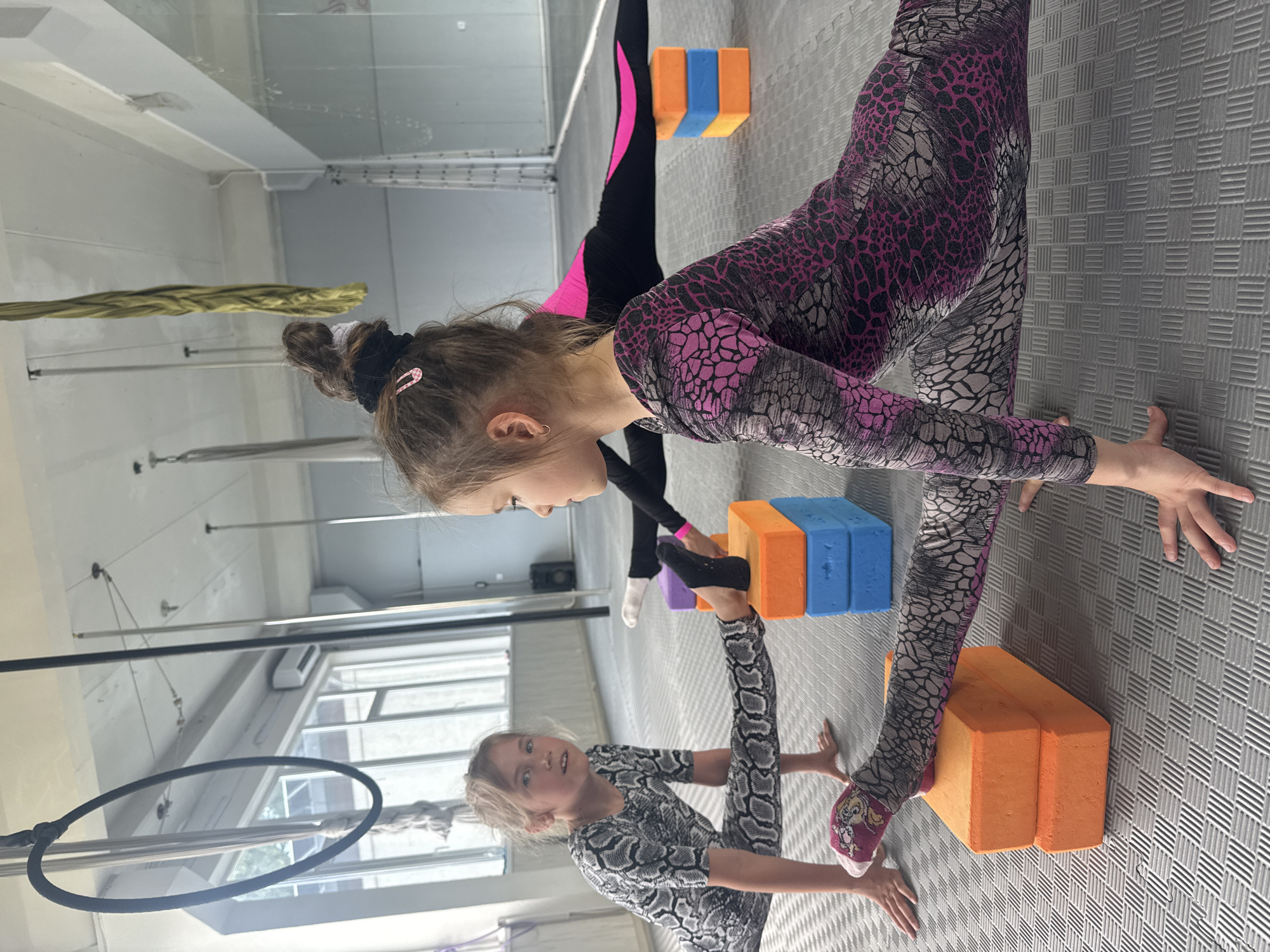 In a well-lit gymnasium, two young girls are engaged in acrobatic training. The girl in the foreground, wearing a black and white patterned outfit and pink-and-white patterned socks, is in a straddle split, with her hands on the floor for support. She looks towards the camera with a serious expression. The second girl, further back and partially obscured, wears a pink and black patterned leotard and black leggings with a pink stripe. She is also in a split, with one leg elevated on a stack of colorful yoga blocks (orange and blue). Her focus appears to be on her stretch, with her body positioned in profile.
The setting is a gym with a grey, textured mat floor. In the background, there are various pieces of gymnastic equipment, including a hoop, poles, and what appear to be aerial silks hanging from the ceiling. Large windows in the background suggest it might be daytime. The overall atmosphere is one of focused athletic activity. There is no visible text in the image.