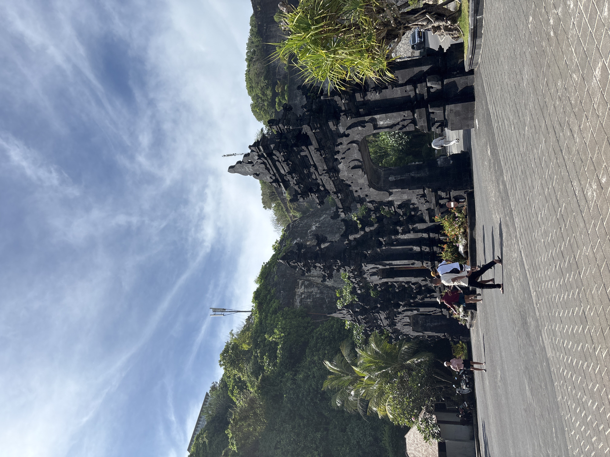 The image depicts the Pura Luhur Uluwatu temple in Kuta Selatan, Bali, Indonesia, from a slightly low angle. The temple, constructed from dark stone, is built into the side of a cliff lush with green vegetation. The sky is partially covered with streaky clouds. Several people are walking on the paved path in front of the temple; a vehicle is also visible in the background.