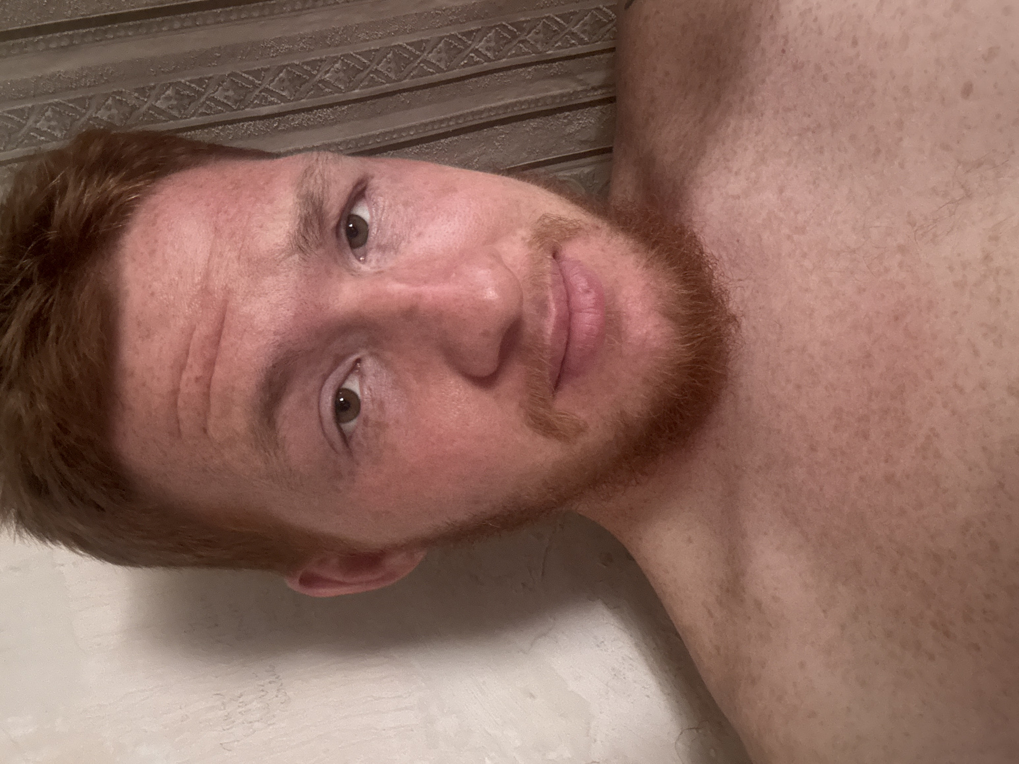 The image is a close-up selfie of a fair-skinned man with short, reddish-brown hair and a light beard and mustache. He has brown eyes and freckles scattered across his face and upper chest. The man is shirtless. The background on the left appears to be a textured, off-white wall with some faint markings. On the right, there is a striped curtain or fabric. The lighting is warm, creating a slightly rosy tone on the man's skin. There is no identifiable location in Makiivka, Ukraine visible in the image.