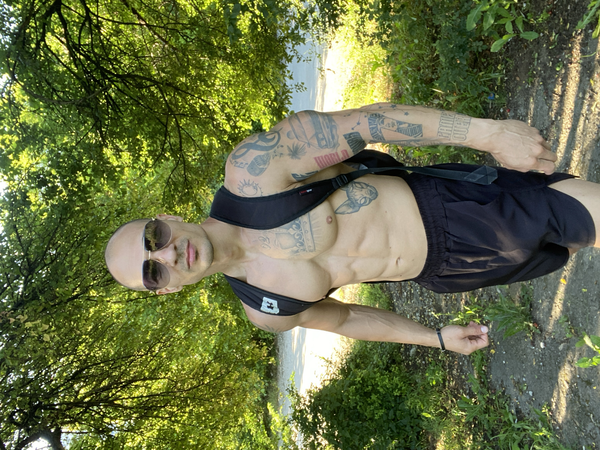 A muscular, bald man with sunglasses and tattoos on his arms and chest stands in a wooded area. He is wearing black athletic shorts and a black backpack. The man is looking directly at the camera. The background consists of trees and foliage, with a dirt path visible. The image appears to have been taken outdoors during the daytime.