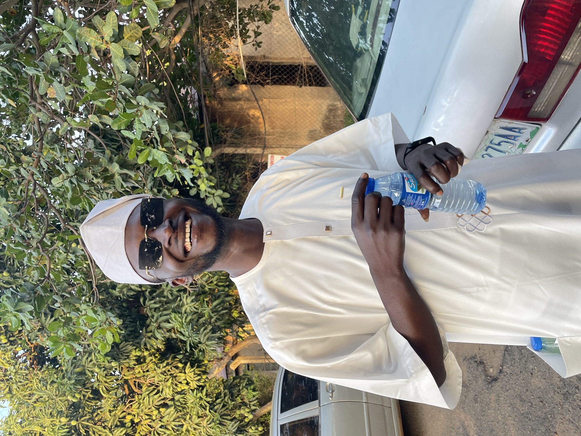 A medium shot depicts a smiling, dark-skinned man holding a water bottle. He wears a white outfit with a matching cap and dark sunglasses with gold accents. A white car is behind him. A license plate that partially reads "275AA" is visible. Green trees are visible around the man. The image may be taken in Jos, Nigeria, as mentioned in the prompt.