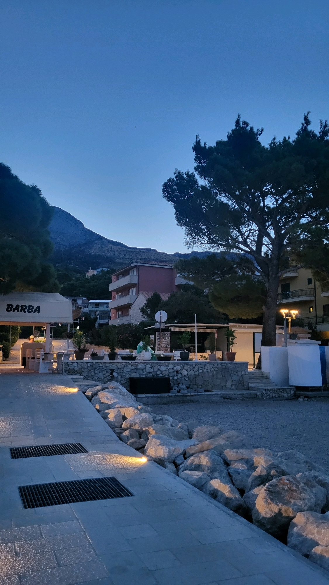 The image depicts a twilight scene along a waterfront promenade in Općina Tučepi, Croatia. The sky is a deep blue, transitioning from the fading daylight. On the left, a white awning with the word "BARBA" printed on it shades an outdoor seating area with tables and chairs. Further back, buildings of various sizes and colors are visible, with a large, dark mountain range forming the backdrop. A prominent, mature tree with dense foliage stands in the mid-right of the frame, its branches casting shadows. In front of the tree, a lamppost with multiple lights illuminates the area. The promenade itself is paved with large, flat stones and features two rectangular grates, possibly for drainage. The edge of the promenade is lined with large, grey rocks, and beyond that, a gravelly area leads to the water, which is not visible. The overall atmosphere is calm and serene, indicative of the early evening. There are no discernible people engaged in activity, though a person is faintly visible sitting at a table under the "BARBA" awning.