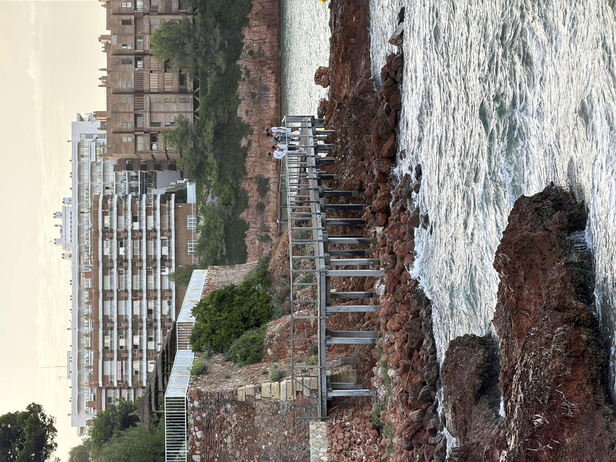 A boardwalk made of weathered wood stretches along a rocky coastline, leading to a pier where four individuals stand observing the sea. The pier is equipped with a metal railing for safety. The coastline is characterized by jagged, reddish-brown rocks and some sparse green vegetation. In the background, several multi-story buildings rise above a dense line of trees. The sky is overcast, suggesting a late afternoon or early morning setting, with soft light illuminating the scene. The water is choppy, with small waves breaking against the rocks. The architecture of the buildings suggests a resort or residential area, consistent with the coastal areas of Salou, Spain.