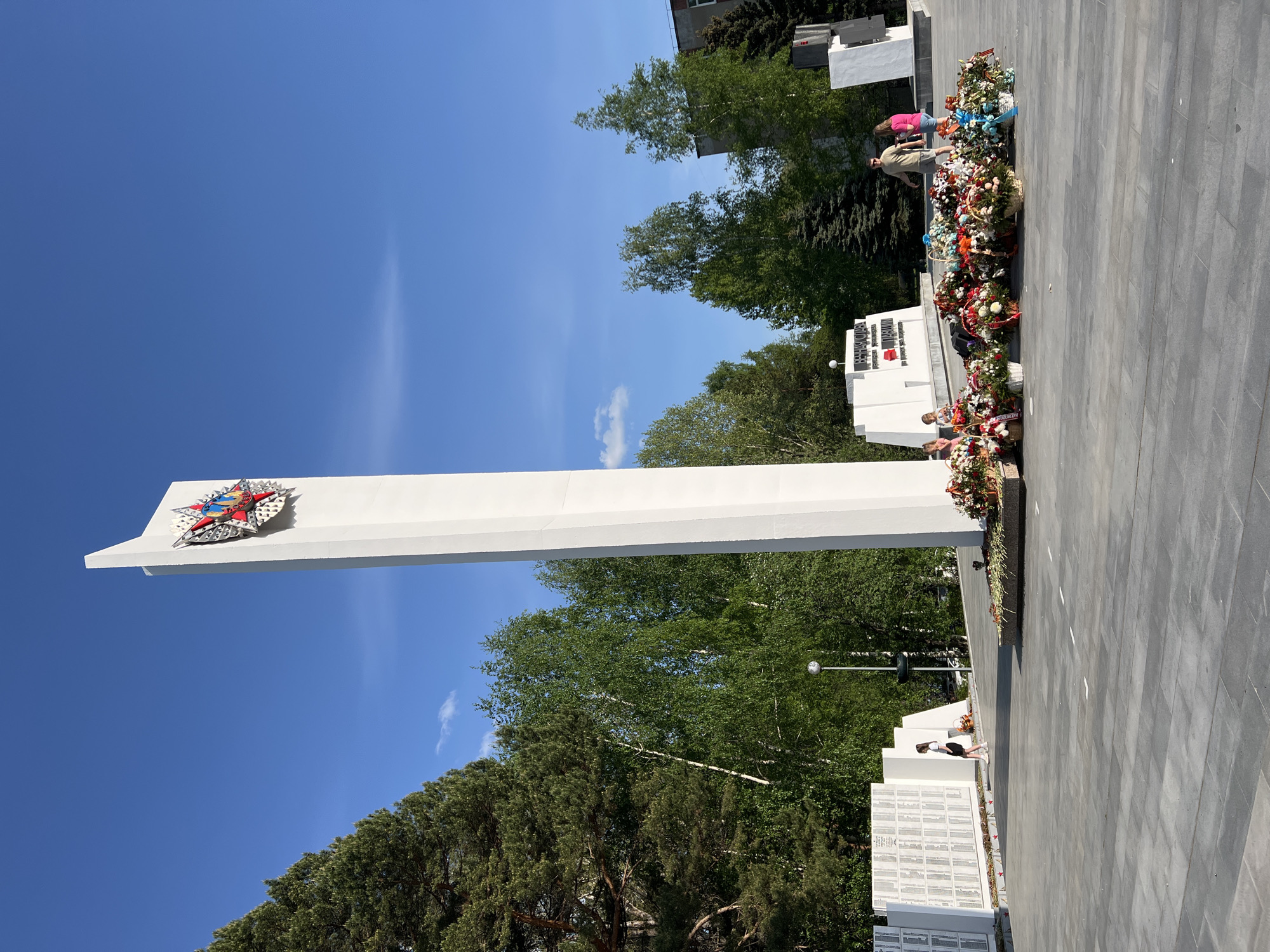 The image shows a white obelisk monument topped with a star emblem, located at a memorial complex in the city of Chaikovsky, Russia. The monument stands tall against a clear blue sky, with green trees visible in the background. Flower wreaths and bouquets adorn the base of the monument and the surrounding area. Several individuals are visible near the monument, including a group of people on the right and a girl looking at memorial plaques on the bottom right. The ground is paved with gray stone tiles.