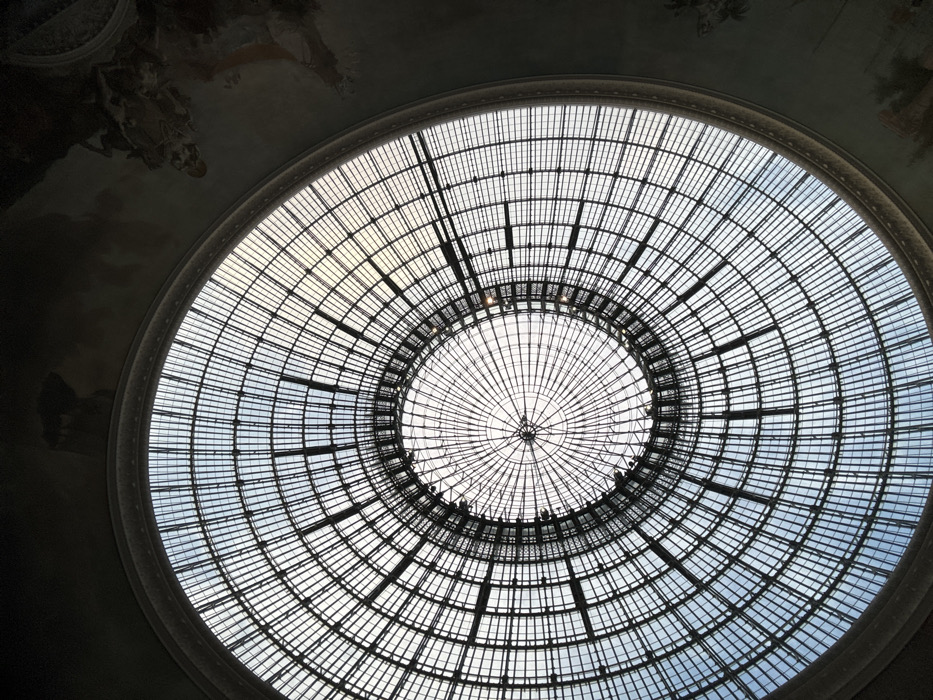The image presents a striking upward view of a grand, circular glass dome or skylight, forming a central feature of an ornate ceiling. The dome is characterized by an intricate structural framework composed of dark, grid-like elements that create a symmetrical pattern of concentric circles and radial spokes. This latticework is prominently silhouetted against the bright sky visible through the glass panels.
Through the transparent sections of the dome, the sky appears bright, with variations in light intensity. Some areas show a clear, light blue sky, while others are diffused, suggesting either varying cloud cover or the way light is filtered by the structure. The innermost section of the dome features a tighter grid of radial lines converging towards the center, emphasizing the circular geometry. Along one of the inner rings of the dome, very small, dark silhouettes can be discerned, which might be decorative elements, lights, or even distant figures, though their specific nature is not clearly identifiable.
Surrounding the brilliant dome, the rest of the ceiling is significantly darker and curved, indicating the overall architecture of the building. This darker area appears to have faint, muted patterns or frescoes, possibly painted scenes or decorative motifs, although they are largely obscured by shadow and the upward viewing angle. The contrast between the brightly lit dome and the dimly lit, decorated ceiling creates a sense of depth and architectural grandeur. The overall impression is one of a magnificent historical building, possibly a museum, gallery, or a grand hall with an elaborate roof structure designed to maximize natural light. No discernible text is visible in the image.