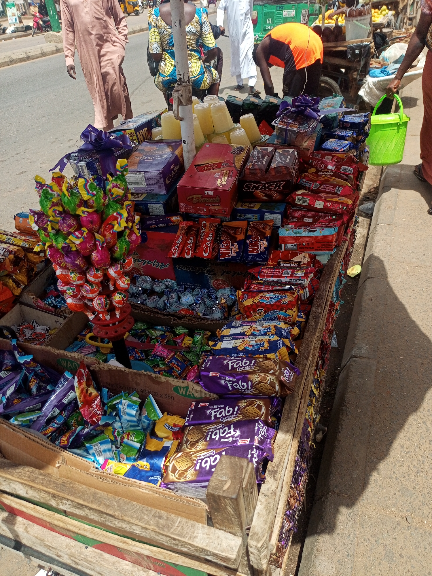 The image shows a street-side confectionery stall in Tempure, Nigeria. The stall, constructed of wood and cardboard, is piled high with various sweets, chocolates, and packaged snacks. Dominating the left side are large numbers of lollipops arranged in a colorful bouquet. Packaged goods include "Fab!" chocolate cookies, "Chairman Snack" items, and various chocolate bars. Containers of a milky-yellow liquid sit near the top of the display. Several people are in the background, including a man in an orange shirt, another in a brown robe, and a woman holding a green bucket. In the far background, traffic and other vendors are visible. The scene is set on a bright, sunny day, casting a shadow on the sidewalk next to the stall.