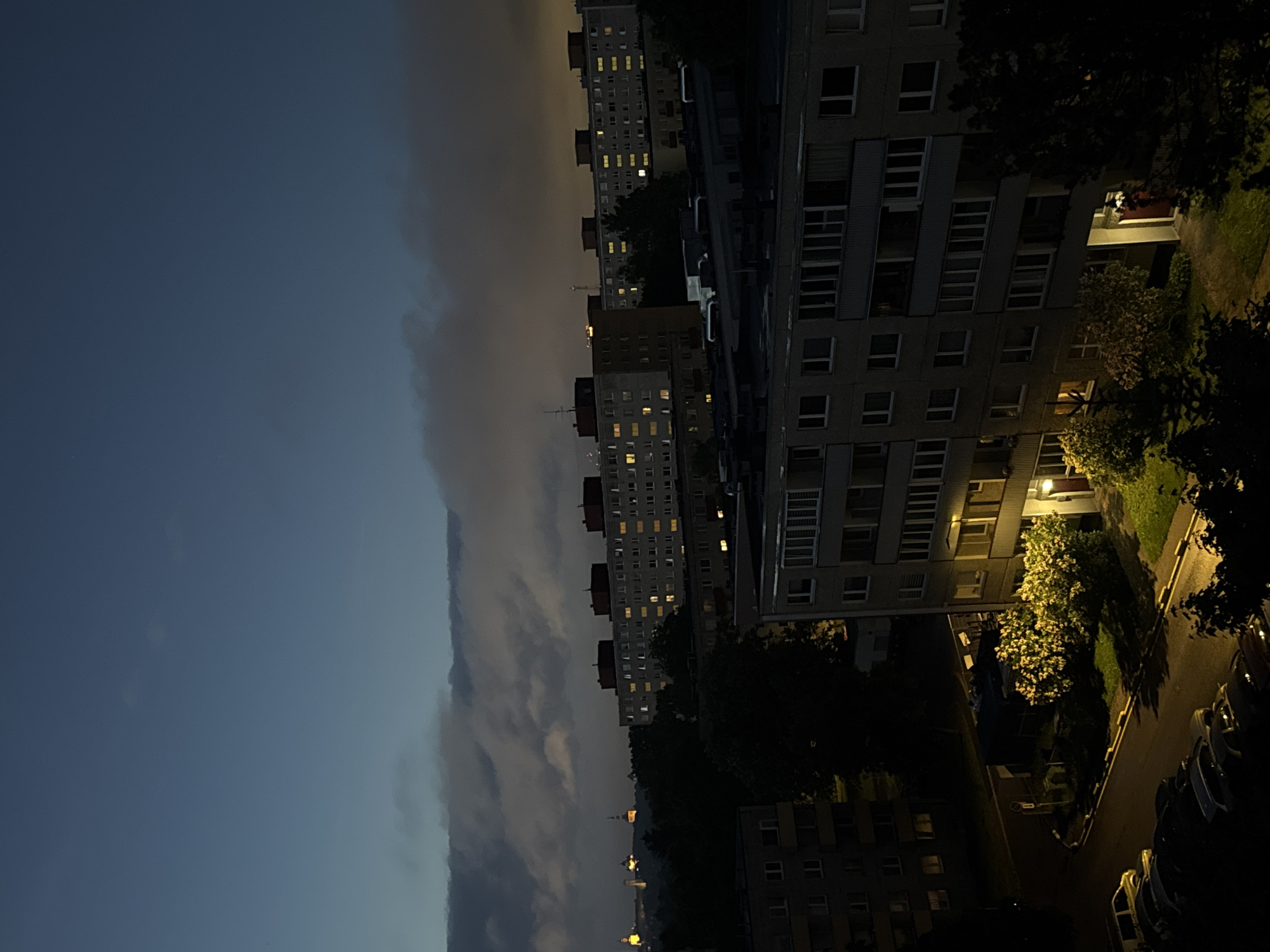 This is an aerial, low-light, horizontal photograph of an urban landscape in Tallinn, Estonia, at dusk or night. The sky is a deep blue, with scattered, illuminated clouds in the middle ground. In the distance, silhouetted by a hazy glow, are several towers and spires that suggest a historical city center, likely Tallinn's Old Town, with its prominent churches and towers. In the mid-ground, a row of apartment buildings stretches across the frame, characterized by their dark facades and illuminated windows, indicating that residents are home. The apartment blocks have multiple stories and balconies. In the foreground, another apartment building, this one closer and larger, dominates the right side of the image. Its windows are also lit, and a bright yellow light illuminates the entrance. A well-maintained grassy area with flowering bushes is visible in front of this building. Adjacent to the building, a street curves away, lined with parked cars. The overall impression is of a residential area of Tallinn, with the older city center visible in the distance, bathed in the soft light of evening.