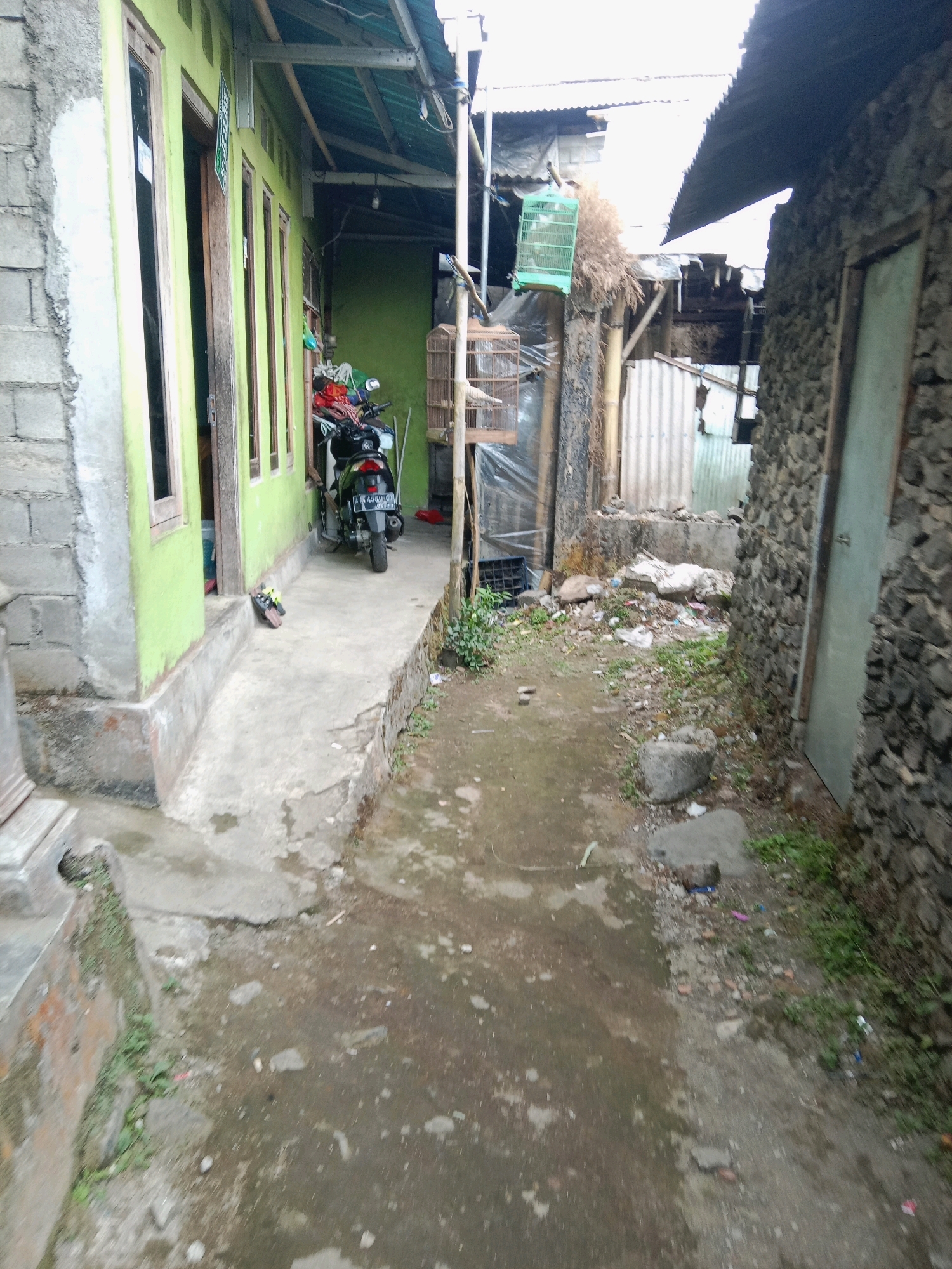 The image depicts a narrow, outdoor alleyway in a residential area of Adipuro, Indonesia. The alley is bordered by two buildings. On the left, a pale green painted house with several windows and an open doorway is visible. A motorcycle is parked on a concrete path in front of this house. On the right, a building constructed with dark, rough-hewn stones forms the alley's boundary, featuring a closed door with a light-colored frame. The ground is a mixture of concrete and dirt, with scattered debris, including plastic bags, stones, and dried vegetation. Several bird cages are hanging from a bamboo structure in the middle ground, suggesting a domestic setting. The overall impression is one of a somewhat unkempt, utilitarian outdoor space within a densely populated area.