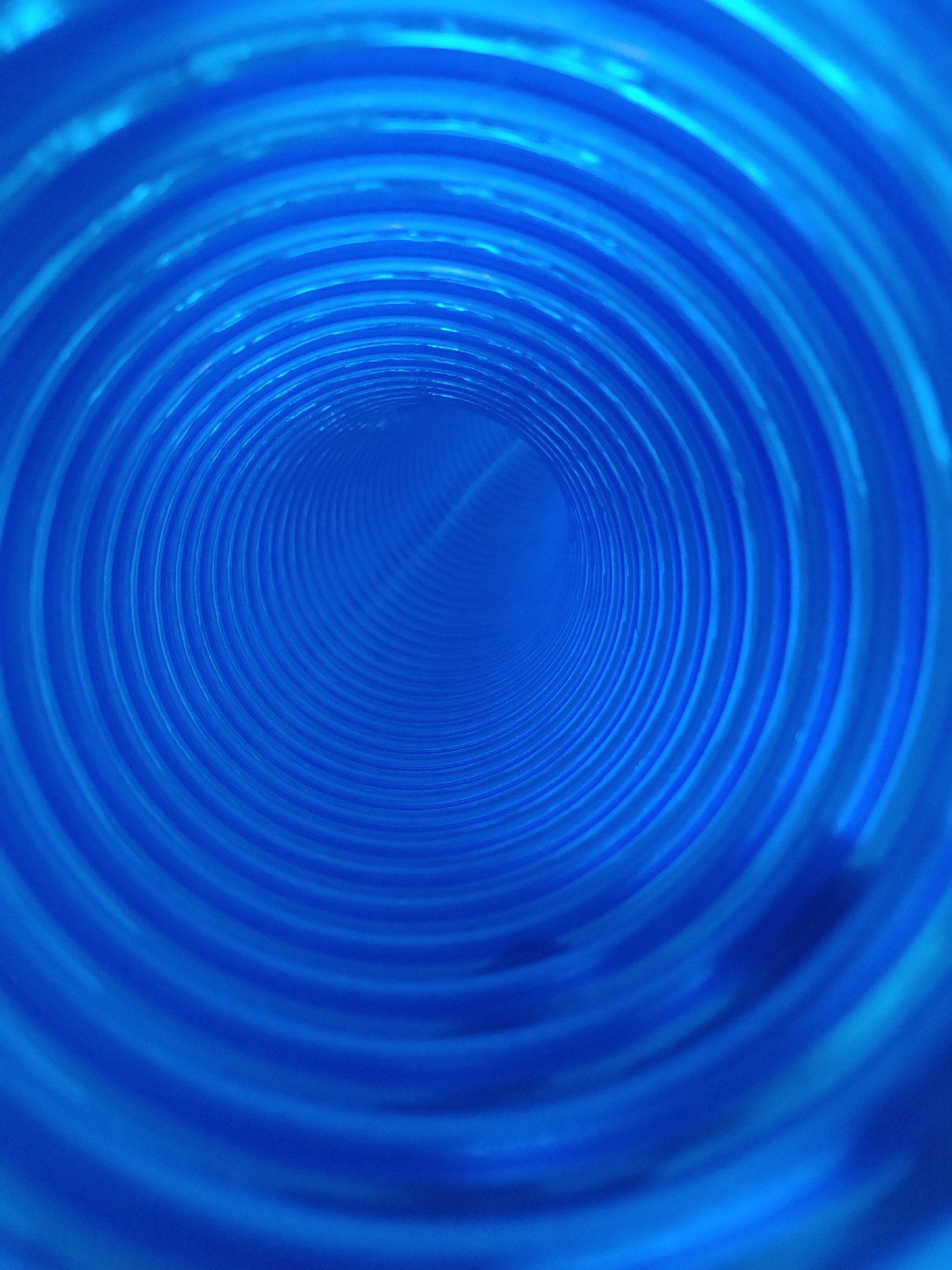 The image presents an abstract view into the interior of a bright, vibrant blue cylindrical structure, resembling a tunnel or a pipe. The perspective is from one end looking directly down its length, creating a strong sense of depth and an optical illusion of endless recession.
The dominant subject is the tunnel itself, characterized by its intensely blue color and corrugated, ribbed inner surface. These ribs form a series of concentric circles that grow progressively smaller and closer together as they approach the distant, central vanishing point. The light source appears to be from the foreground, illuminating the interior and causing subtle gradations in the blue hue, with lighter, brighter areas near the entrance and a slightly darker, more muted blue towards the vanishing point. Highlights are visible along the ridges of the corrugations, emphasizing the texture.
There are no people, objects other than the tunnel structure, or specific events taking place. The scene is static and devoid of discernible activity. No specific time of day, weather conditions, or emotional states can be inferred from the image. There are no visible location cues within the image itself, nor is any text present. The overall impression is one of pattern, color, and profound depth. The provided location context is Meunasahaluedrienlhoksukon Barat, Indonesia.