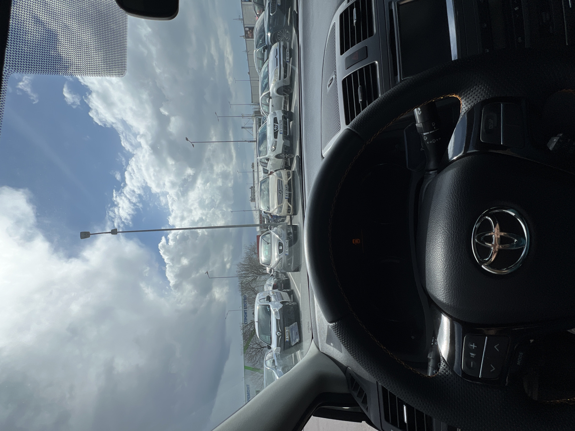 The image presents a view from the driver's seat of a Toyota vehicle, featuring a black perforated leather steering wheel with orange stitching and an illuminated orange warning indicator on the instrument cluster. The vehicle is stationary in a large outdoor parking lot.
Numerous other vehicles are parked in the midground, including a white Renault Kangoo van with Bulgarian license plate B 5226 XB, a grey SEAT with license plate A 4221 HC, a light-colored Mercedes-Benz with license plate A 6526 HT, a white Nissan Qashqai with license plate A 2316 PA, and a silver Citroën C4 with license plate A 3597 MA. Multiple tall, grey streetlights are visible throughout the parking area.
In the background, a large, light-colored commercial building with a horizontal green stripe displays Cyrillic text, including "СТРОИТЕЛСТВО" (Construction) and "РЕМОНТ" (Repair/Renovation). Bare deciduous trees are situated between the parking lot and the building. The sky is partially cloudy with patches of blue visible.
Based on the Bulgarian license plates (starting with 'B' and 'A') and Cyrillic signage, the location is identified as Burgas, Bulgaria. The building's text suggests a large construction or home improvement store, typical of a retail park environment in the city.
