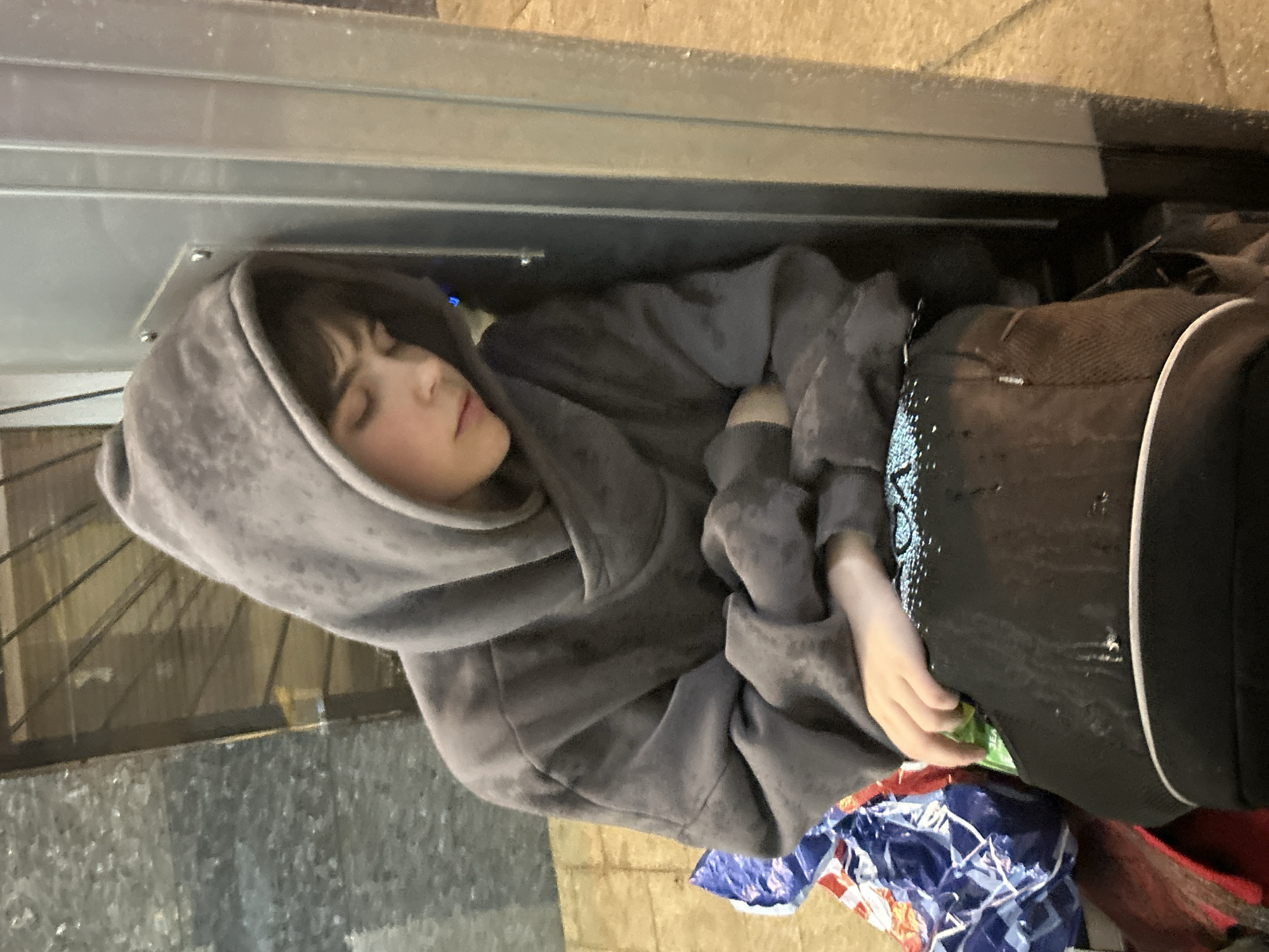 A young person wearing a grey hooded sweatshirt is resting their head against a metal pole, eyes closed, in an urban environment. They are seated on a black bag with white trim and what appears to be a blue and white patterned bag to their left. The ground is paved with light-colored tiles. The background shows a textured grey wall and a darker, possibly metallic structure with visible bars. The lighting suggests it is nighttime or an indoor, dimly lit area.