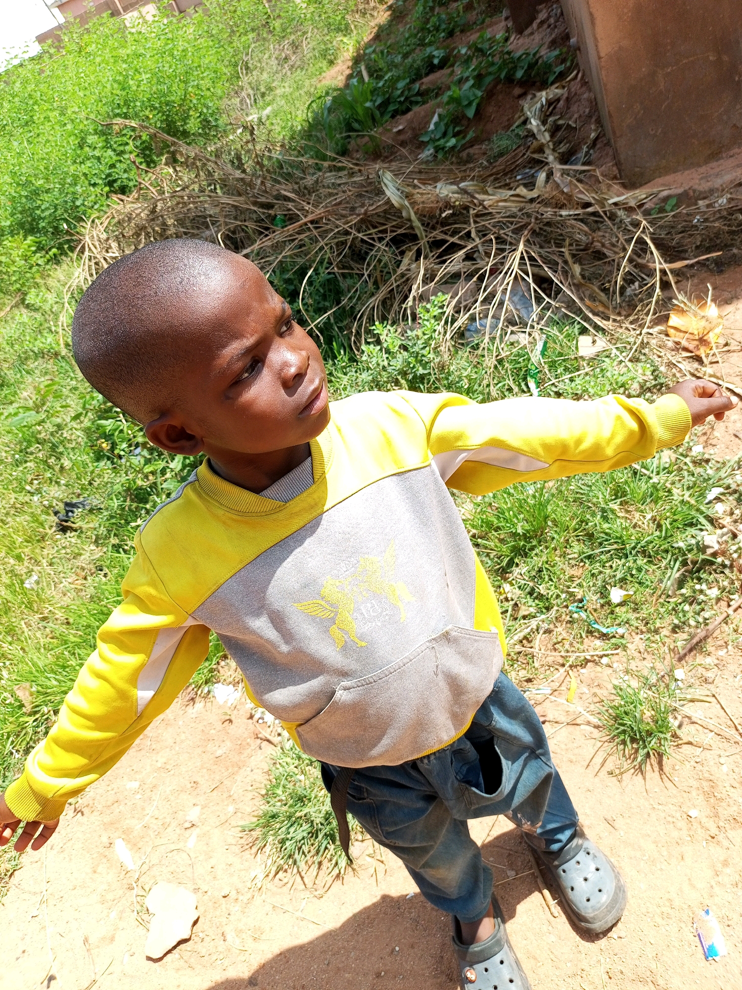 A young Black male, appearing to be around 5-7 years old, stands in a dirt and grass area under a bright sun. He is facing away from the camera, looking to his right with his head tilted slightly up. He has short, dark hair and dark skin. He is wearing a yellow and gray long-sleeved shirt with a kangaroo pocket and dark blue jeans. On his feet, he wears dark gray slip-on clogs. His left arm is extended to his left, and his right arm is bent, with his hand pointing to his right. The ground is a mix of reddish-brown dirt and green grass, with scattered dry twigs and leaves. In the background, there is more dense green vegetation and a portion of a reddish-brown structure, possibly a wall or building. The overall setting appears to be outdoors in a rural or semi-urban area.