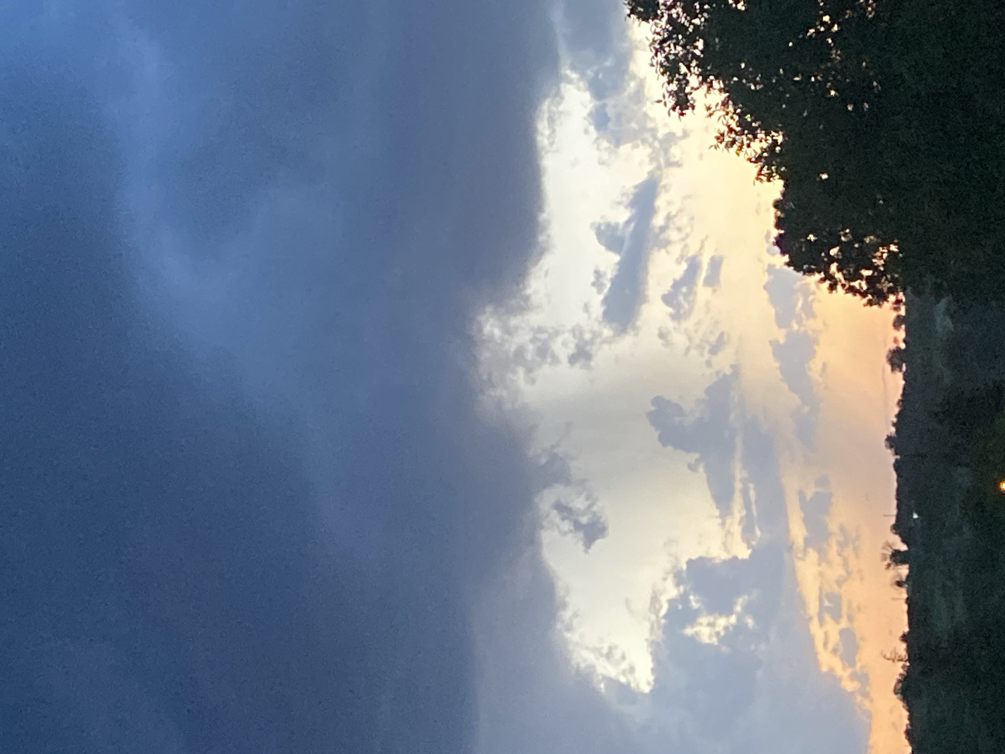 The image shows a cloudy sky. The left side of the image is dominated by a large, dark gray cloud mass. The right side features lighter, illuminated clouds interspersed with hints of orange and yellow, possibly suggesting a sunset or sunrise. Silhouetted tree foliage is visible at the very right edge of the image. There are no people visible in the image. The location is not confirmed.