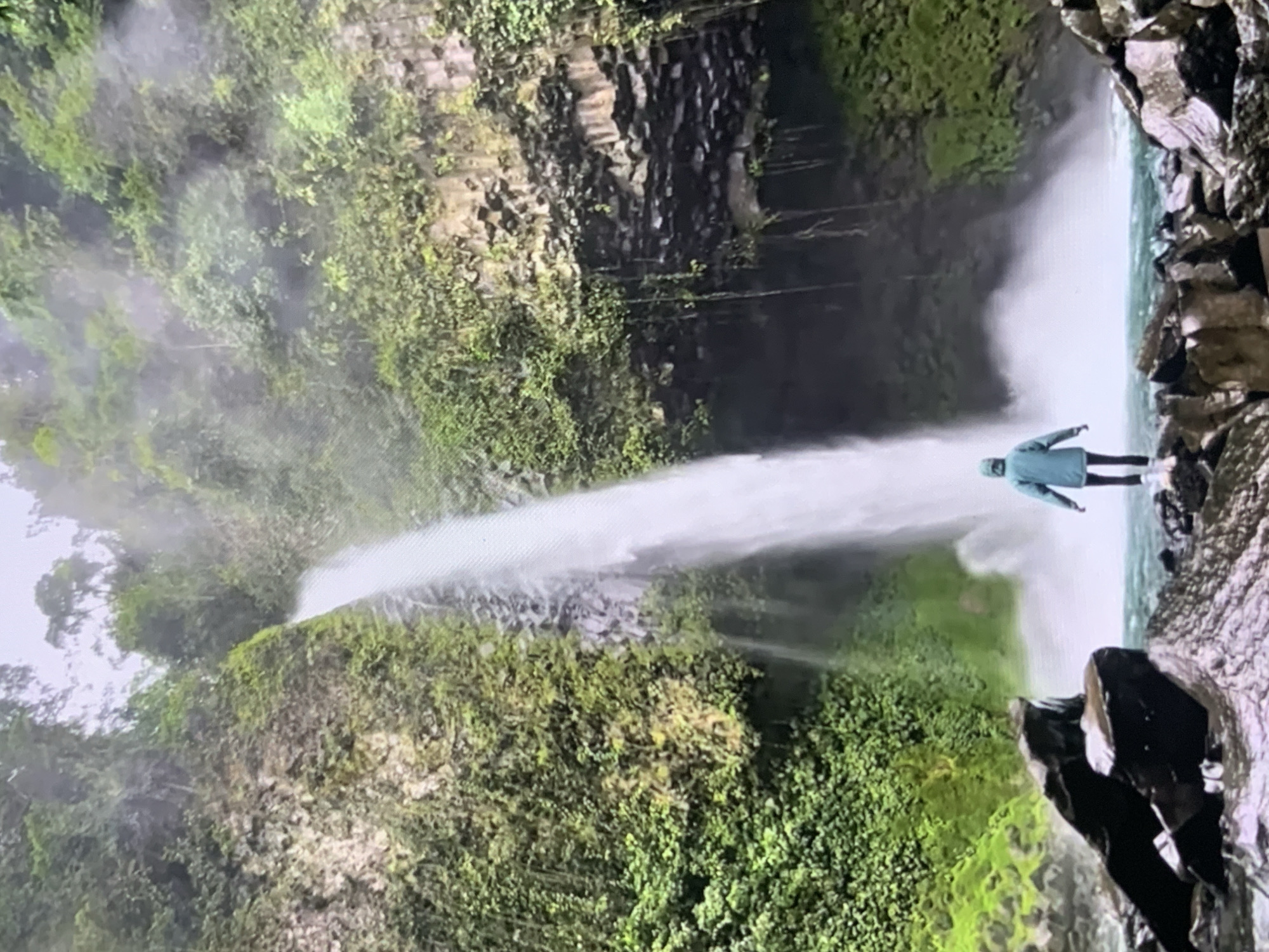 The image depicts a person standing on rocks in front of a large waterfall. The person is wearing a teal hooded jacket and dark pants. The waterfall cascades down a steep, rocky cliff face covered in lush green vegetation. The mist from the waterfall obscures the upper portion of the scene, creating a hazy atmosphere. The foreground consists of dark, wet rocks, and the water at the base of the waterfall is a vibrant turquoise color. The environment appears to be a natural, tropical setting. The image does not show any recognizable landmarks or features specific to the city of Stavropol, Russia.