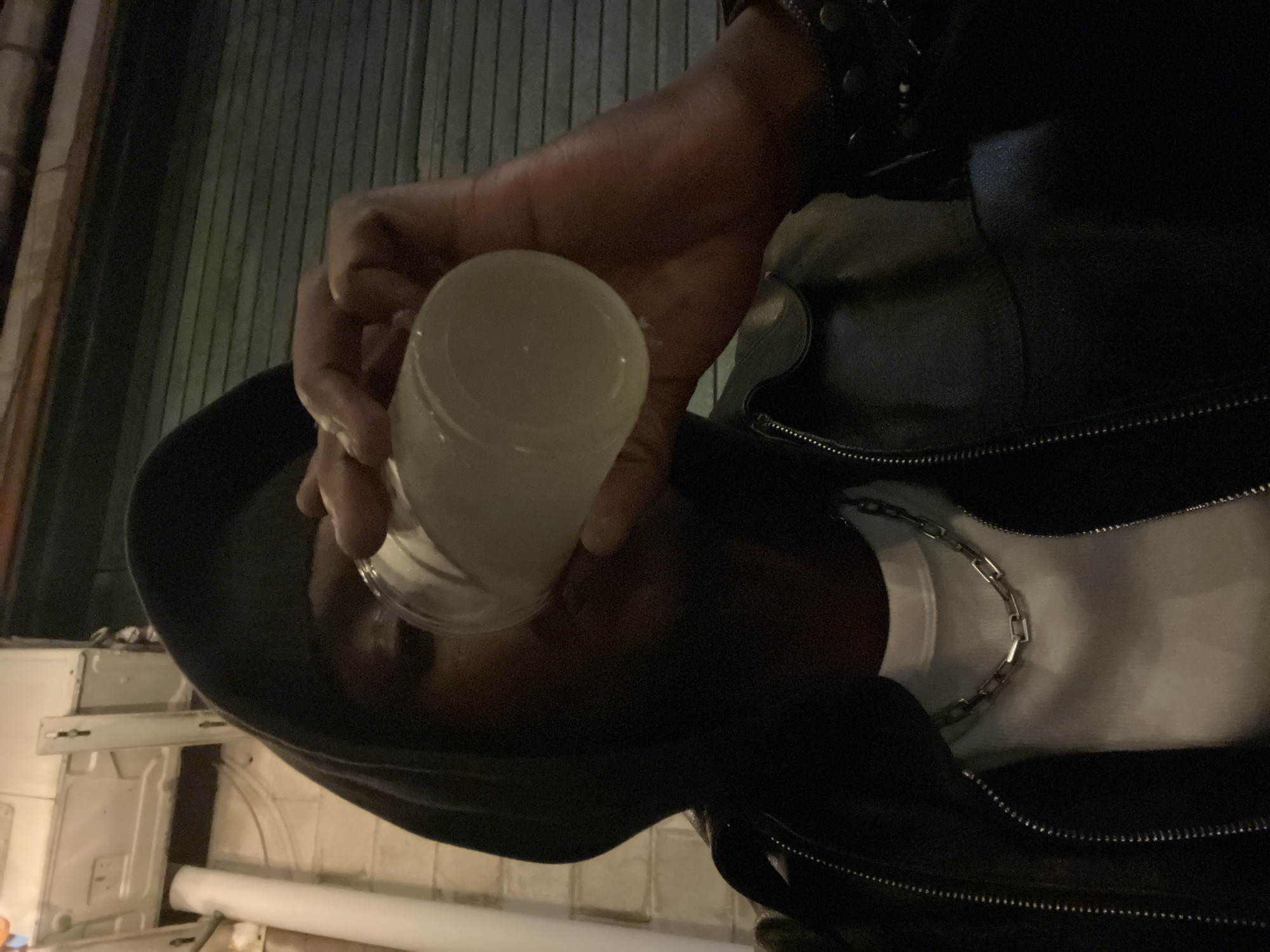 A person wearing a black hooded jacket, a white shirt with a silver chain necklace, and a black leather jacket with a studded bracelet on their wrist is holding a white cup. The background appears to be an urban exterior with grooved metal paneling. Some internal elements are visible in the lower left of the image. There is no specific location in Barcelona that can be determined from this image alone.