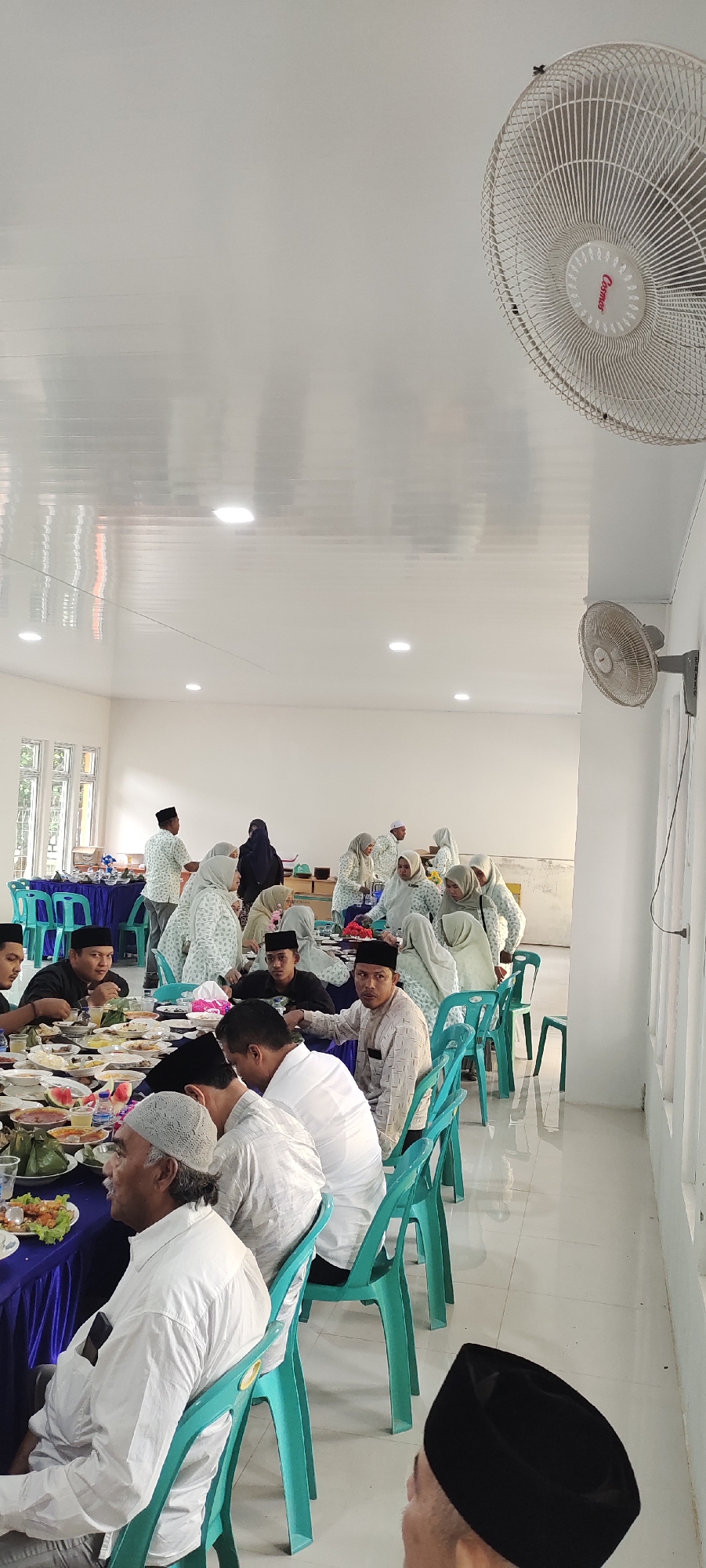 The image depicts an indoor gathering with a meal being served. The scene appears to be a community event or a religious observance, possibly in Peurupok, Indonesia, given the context. A long table is laden with various dishes, and numerous individuals are seated around it, engaged in eating and conversation. Many of the men are wearing white shirts and some have on peci caps, traditional Islamic headwear. Several women are also present, some wearing headscarves. The room is brightly lit by fluorescent lights on a white, corrugated ceiling. A ceiling fan is visible in the upper right corner, and a wall-mounted fan is on the right side of the image. Green plastic chairs are arranged around the tables. The overall atmosphere suggests a communal meal and social gathering.