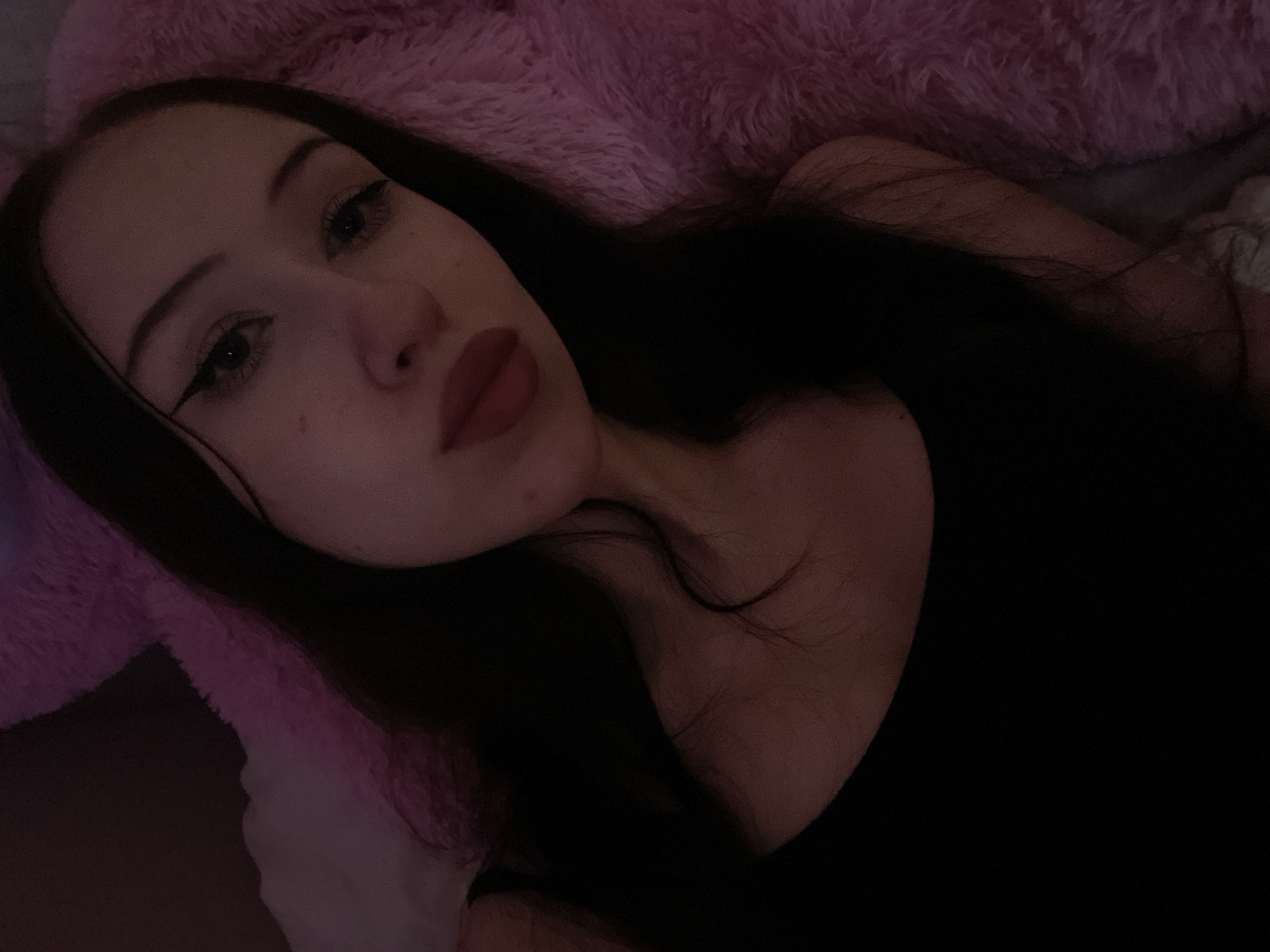 A young woman with long dark hair is captured in a close-up selfie. She is wearing a black tank top and has winged eyeliner. The background is blurred, but appears to be a soft pink, possibly a blanket or a stuffed animal. The lighting is dim and casts a pinkish hue over the scene. There is no indication of the location in Votkinsk, Russia.