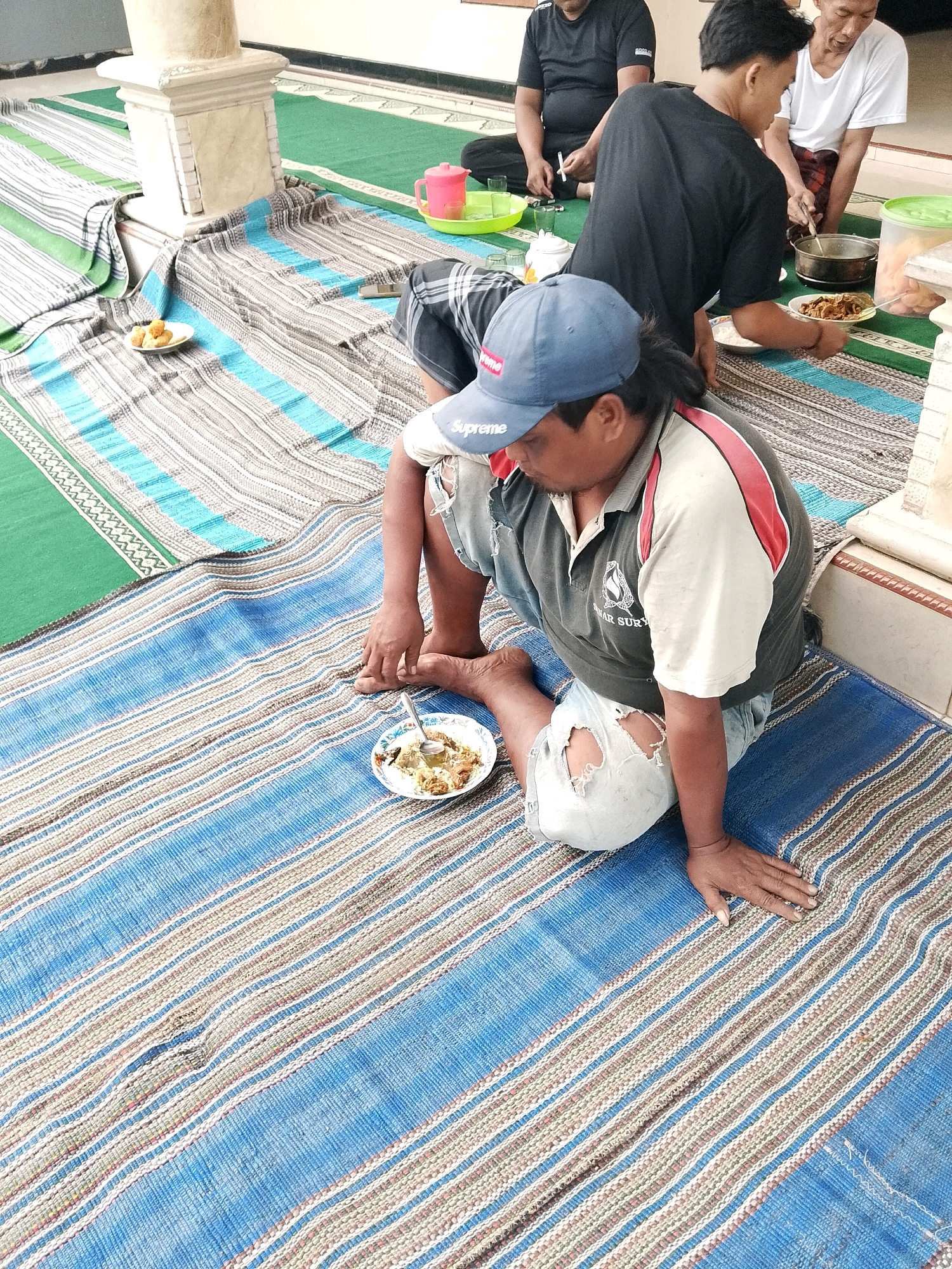 The image depicts an outdoor gathering at a location that appears to be in Karangsari, Indonesia, possibly in a courtyard or open-air area. The ground is covered with woven mats, some in green and others with blue and brown stripes. A group of men are seated on the mats, engaged in eating.
In the foreground, a man wearing a blue baseball cap and a tattered denim shirt is seated on a blue and brown striped mat, looking down at a plate of food in front of him. He has a spoon in the plate. To his left, a small plate with a few fried snacks is visible on a mat.
Behind him, another man in a black t-shirt is serving food from a pot into a white bowl. He is using a ladle. Next to him, a man in a white t-shirt and a sarong is also present, and there's another man in a black t-shirt seated further back on a green mat. Several plastic cups and containers are scattered around, suggesting a meal is in progress. The setting has architectural elements, including a pillar and a white wall, indicating it is likely a residential or community outdoor space.