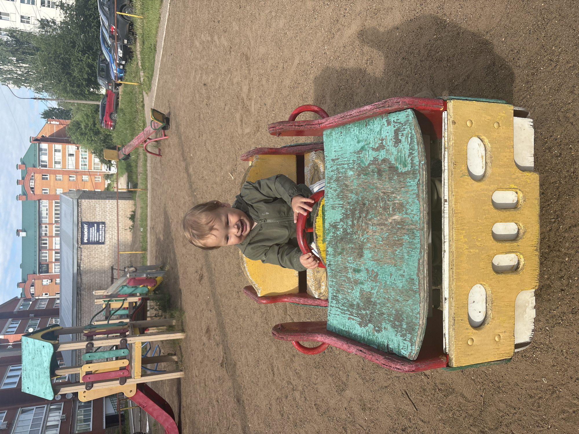 A joyful young child with light brown hair, wearing a green jacket, is depicted playing on a weathered, multi-colored wooden play structure on a sandy playground. The child is seated in the structure, which resembles a vehicle like a car or train, holding a red steering wheel and laughing with a wide, open-mouthed smile. The play vehicle shows signs of age with peeling paint in teal, yellow, red, and white.
The playground itself has a light brown, sandy or dirt ground. Other playground equipment is visible, including a red and yellow seesaw in the mid-ground and a larger, multi-component playset with a slide, steps, and platforms, also made of painted wood in colors like red, yellow, green, and light blue.
In the background, several parked cars (red, blue, black) are visible on a grassy strip next to a paved road. Behind them stand multi-story residential buildings with brick facades and some green roofs, indicative of a residential area in Chaikovsky, Russia. A large black sign on a brick wall features Cyrillic text, with "РЕМОНТ" (REPAIR) at the top and "СЕРВИСНЫЙ ЦЕНТР" (SERVICE CENTER) below, along with contact details. The scene is illuminated by bright daylight under a clear sky, casting noticeable shadows. The overall impression is one of happy childhood play in a communal outdoor space.