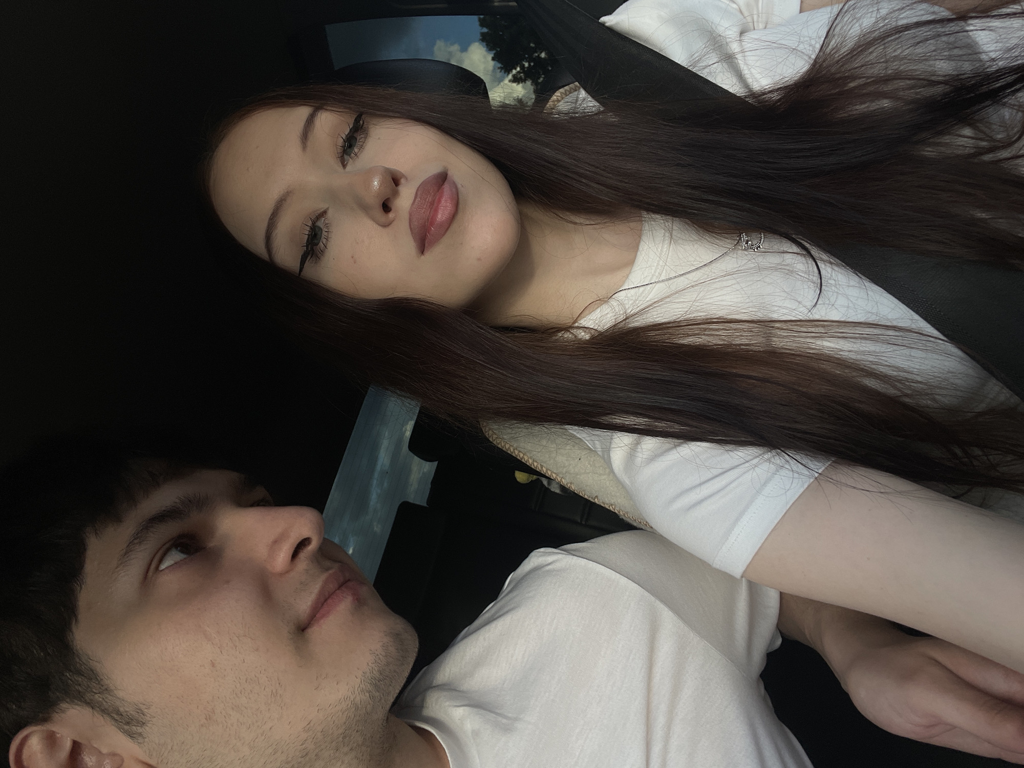 This media file shows a close-up selfie taken inside a car, likely in Gavrilovka, Russia. The photo features a young woman and a young man. The woman is in the foreground, her face angled towards the camera with her eyes looking upwards. She has long, dark brown hair, which is spread across her shoulder and chest. She is wearing a white t-shirt with a subtle neckline detail. Her makeup includes winged eyeliner and lip gloss. The man is in the lower left portion of the frame, looking upwards and away from the camera. He has short, dark hair and is wearing a white t-shirt. The setting appears to be daytime, with natural light filtering into the car, suggesting it could be a sunny or overcast day. The interior of the car is visible, including a seatbelt and the car's interior trim. The overall mood of the photo is intimate and candid. There is no visible text in the image.