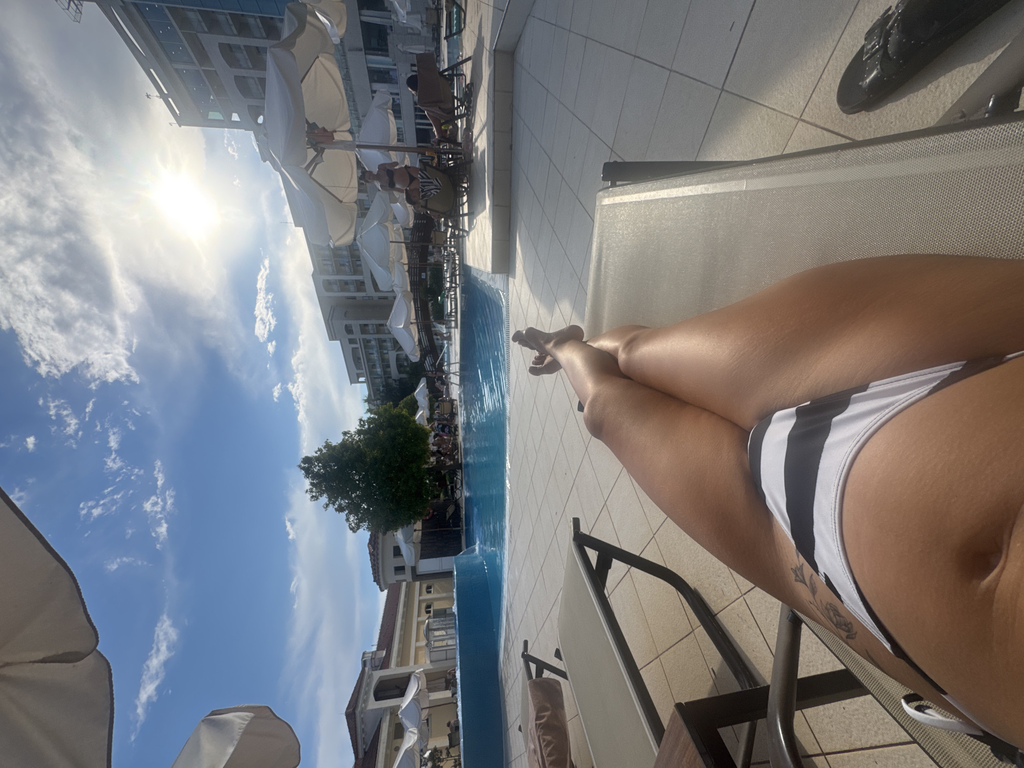 In the city of Boreti, Montenegro, an individual is reclining on a lounge chair beside a swimming pool. The person, partially visible from the waist down, is wearing a black and white striped bikini bottom. Their legs are crossed, and a tattoo is visible on their left thigh. The environment is a poolside area with a tiled patio, lounge chairs, and umbrellas providing shade. In the background, the blue water of the swimming pool is visible, along with a building and trees under a bright, sunny sky with scattered clouds. Several other people are scattered around the pool area, some sitting under umbrellas and others near the water.
