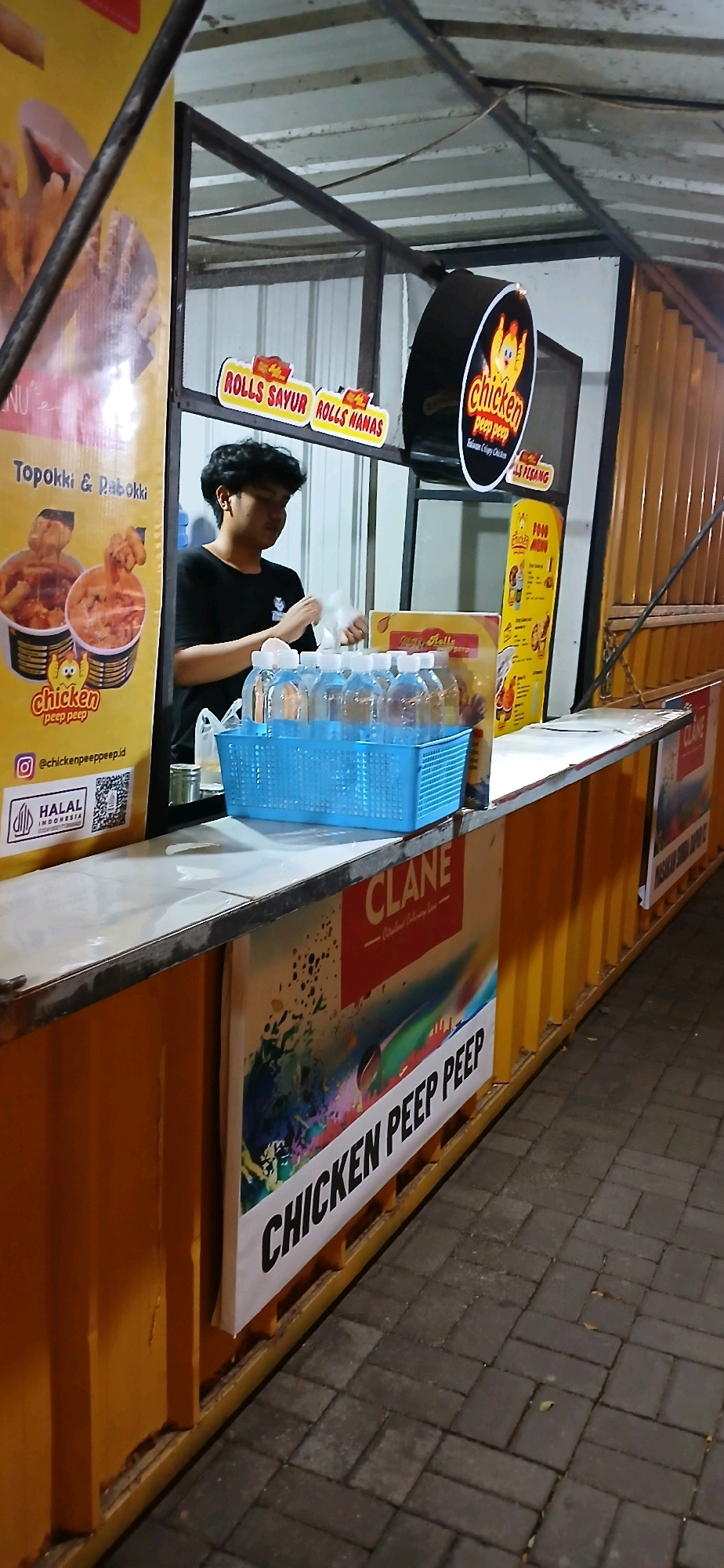 A young male food vendor is seen at a street-side food stall in Cirebon, Indonesia. He is wearing a black t-shirt and appears to be preparing food or packaging an order, as he is holding a white plastic bag. In front of him, on a counter, is a blue plastic basket filled with bottled water. Behind him, the stall has various signs, including one with a cartoon chicken logo that reads "Chicken Peep Peep" and another advertising "Rolls Sayur" and "Rolls Nanas." To the left of the stall, a large yellow sign displays menu items and images of food. The stall itself is made of corrugated metal, painted orange. The ground is paved with dark gray interlocking bricks. A large banner with colorful abstract art and the text "CHICKEN PEEP PEEP" is affixed to the front of the stall. The overall scene suggests a busy outdoor food establishment.