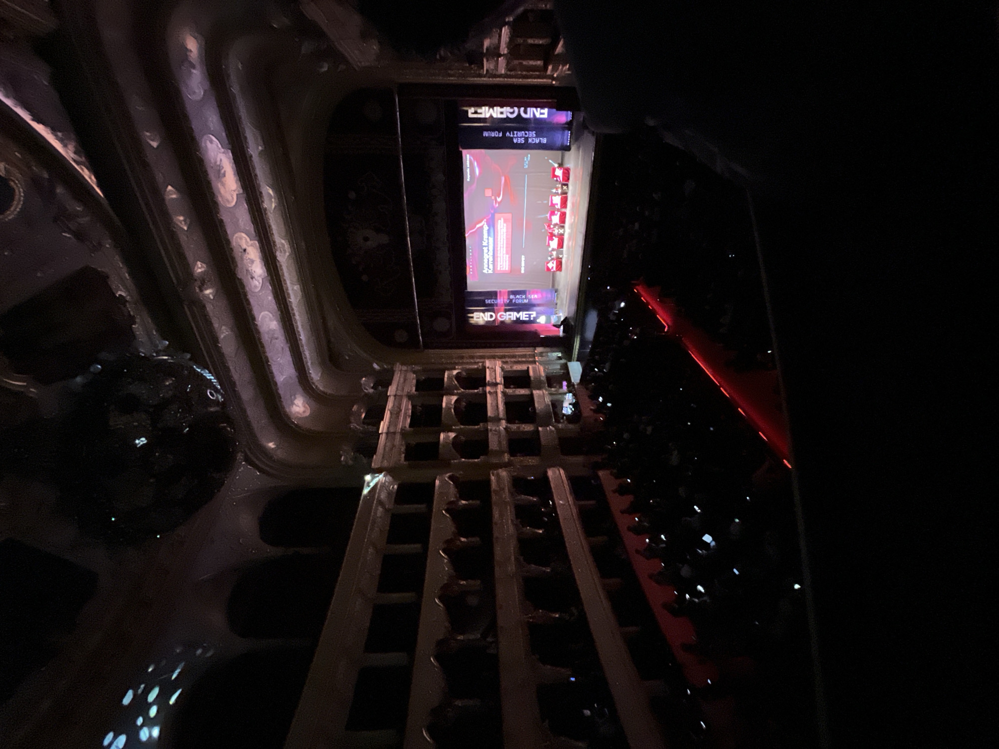 The image shows a view from an elevated position, likely a balcony, overlooking an event. Below is a grid-like structure of some sort. In the distance, a screen displays the words "END GAME?" along with "BLACK SEA SECURITY FORUM". Seated figures are visible below and to the right of the screen, facing it. Red lighting illuminates a section of the seated area. The architecture surrounding the viewing position includes detailed moldings. Based on the text on the screen, this event is likely taking place at a security forum, possibly in Odesa, Ukraine.
